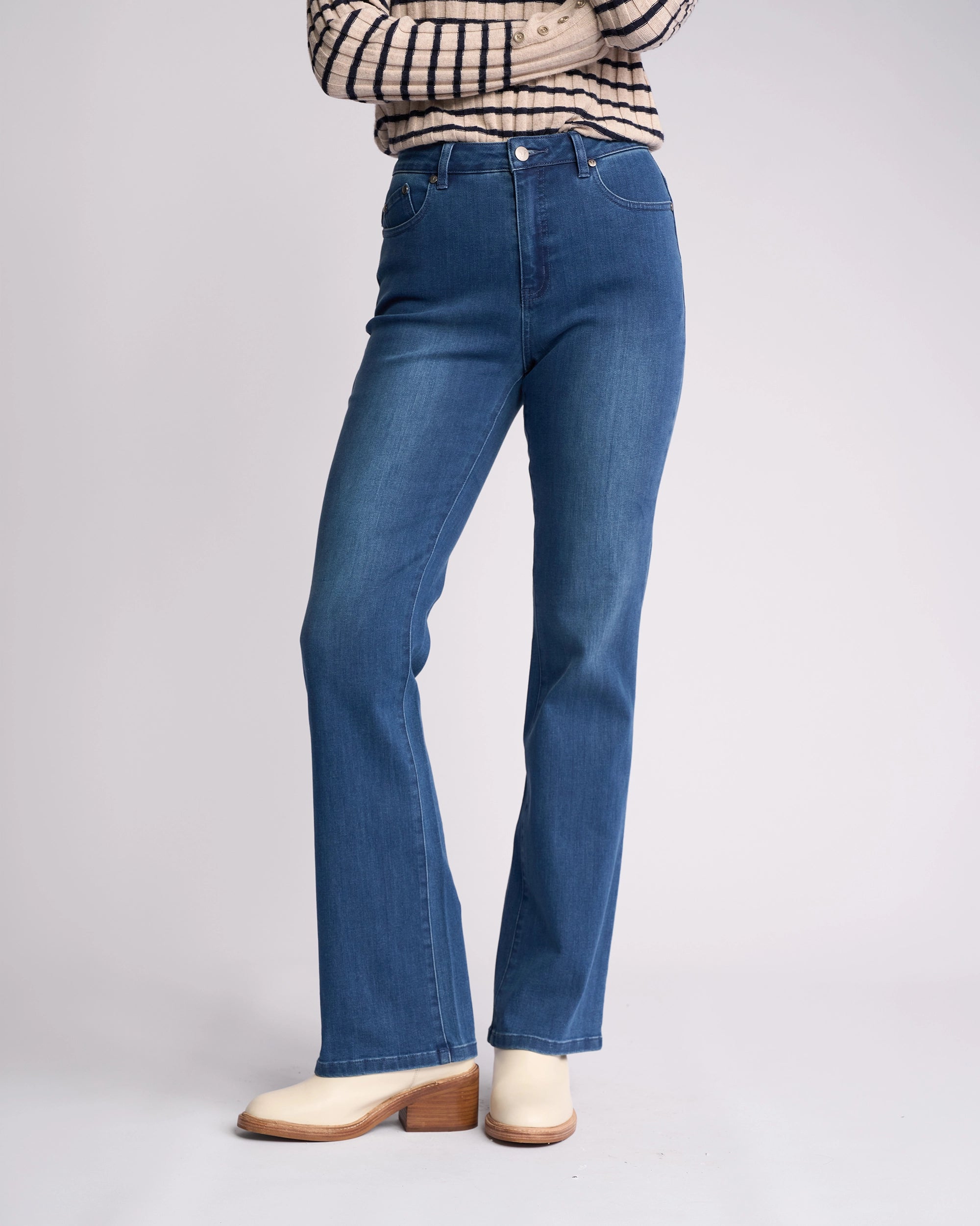 A person wearing Vassalli's Theory Jean - Blue Denim (With Fade), a beige and black striped long-sleeve top, and light-colored heels stands with arms crossed against a plain background.