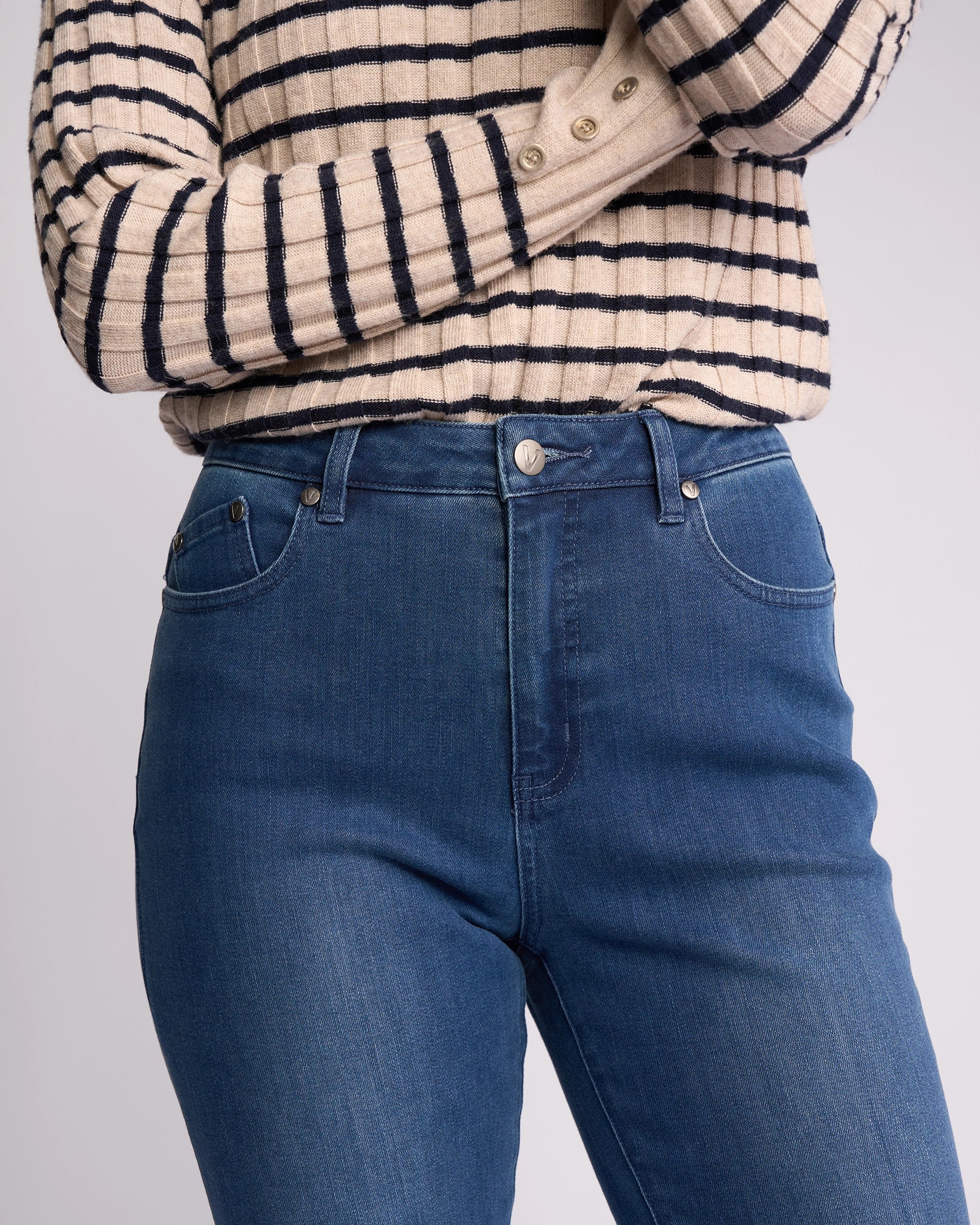 A person, arms crossed and face not visible, stands against a neutral background wearing high-rise Vassalli Theory Jean - Blue Denim (With Fade) and a beige and black long-sleeve striped sweater.