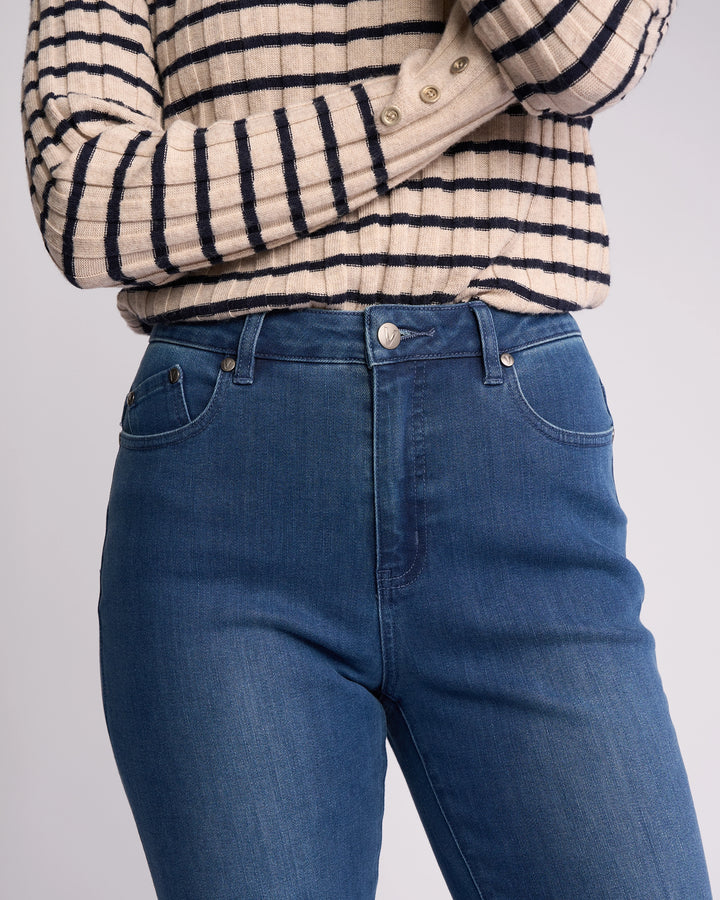 A person, arms crossed and face not visible, stands against a neutral background wearing high-rise Vassalli Theory Jean - Blue Denim (With Fade) and a beige and black long-sleeve striped sweater.