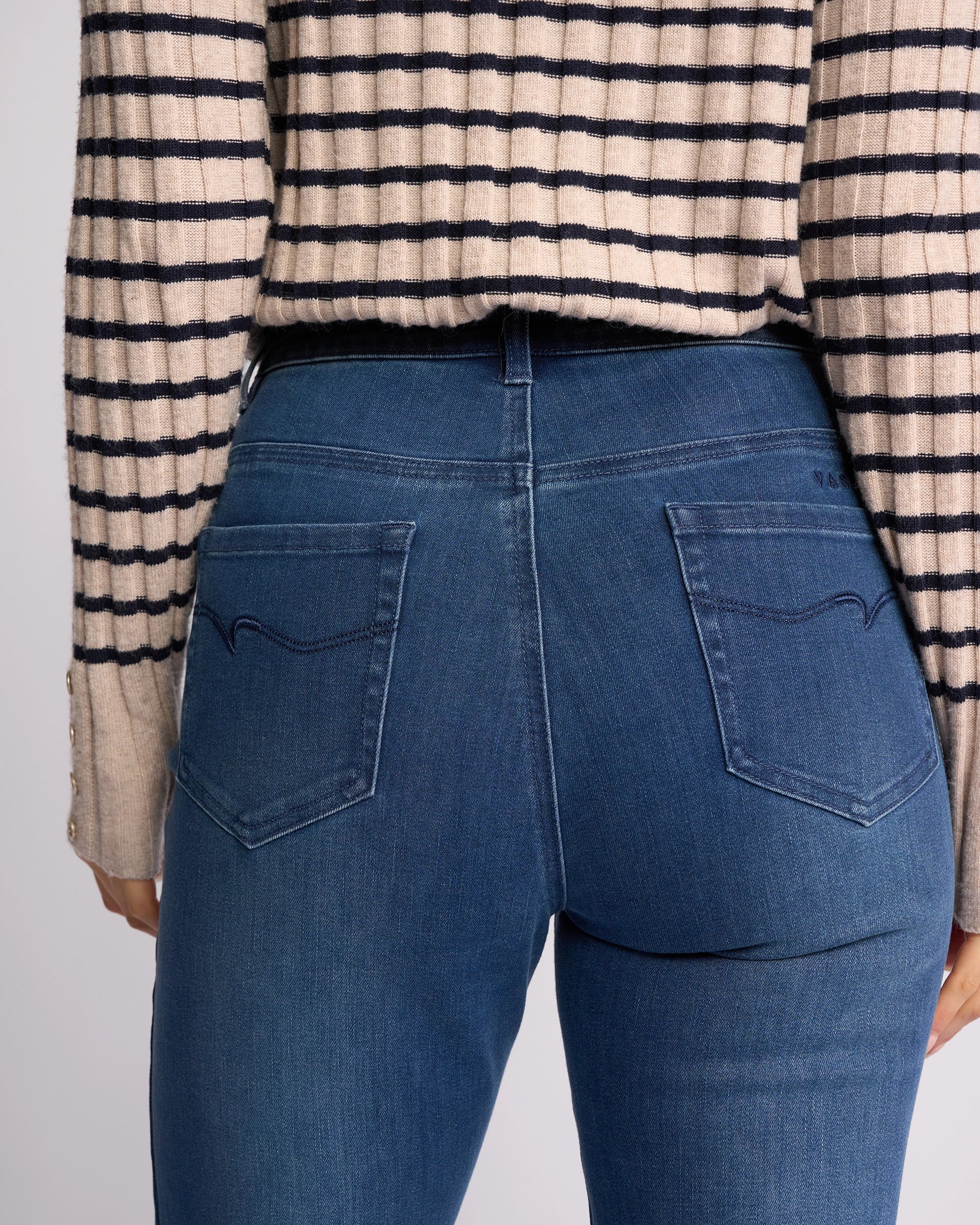 A person is shown from the back wearing Vassalli's Theory Jean - Blue Denim (With Fade) and a beige and black striped long-sleeve top. The jeans have subtle fading, two back pockets, and refined stitching at the waist and upper legs.