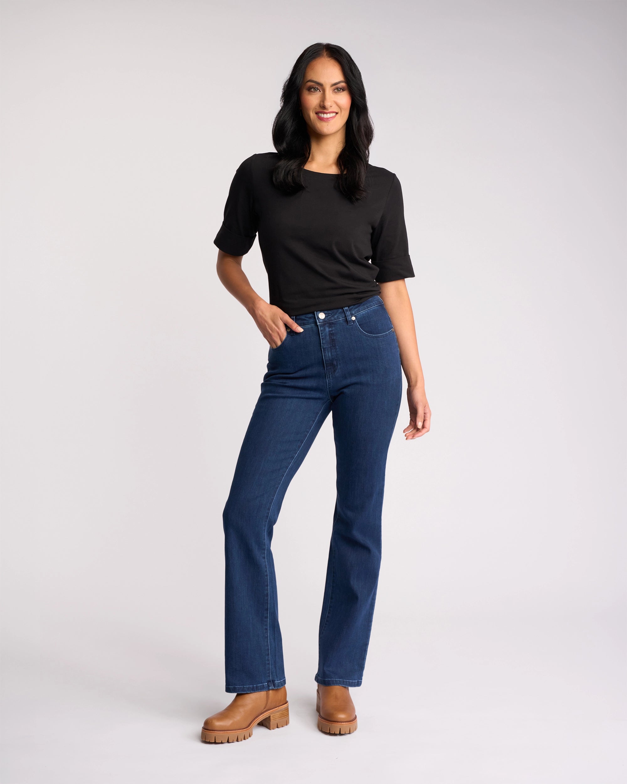 A person with long dark hair, black t-shirt, and tan boots stands smiling with one hand on their hip, wearing Vassalli’s Theory Jean - Blue Denim (Without Fade), against a plain light background.