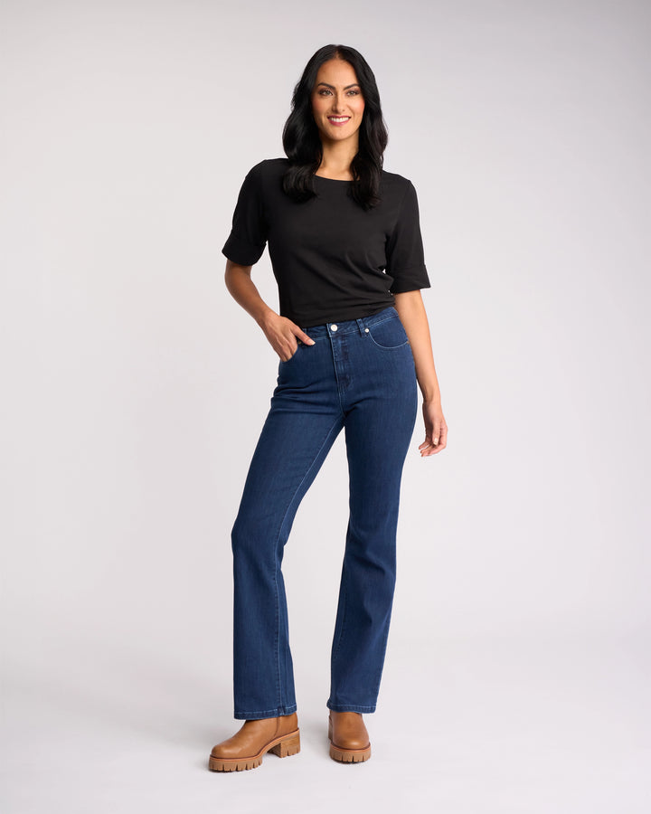 A person with long dark hair, black t-shirt, and tan boots stands smiling with one hand on their hip, wearing Vassalli’s Theory Jean - Blue Denim (Without Fade), against a plain light background.