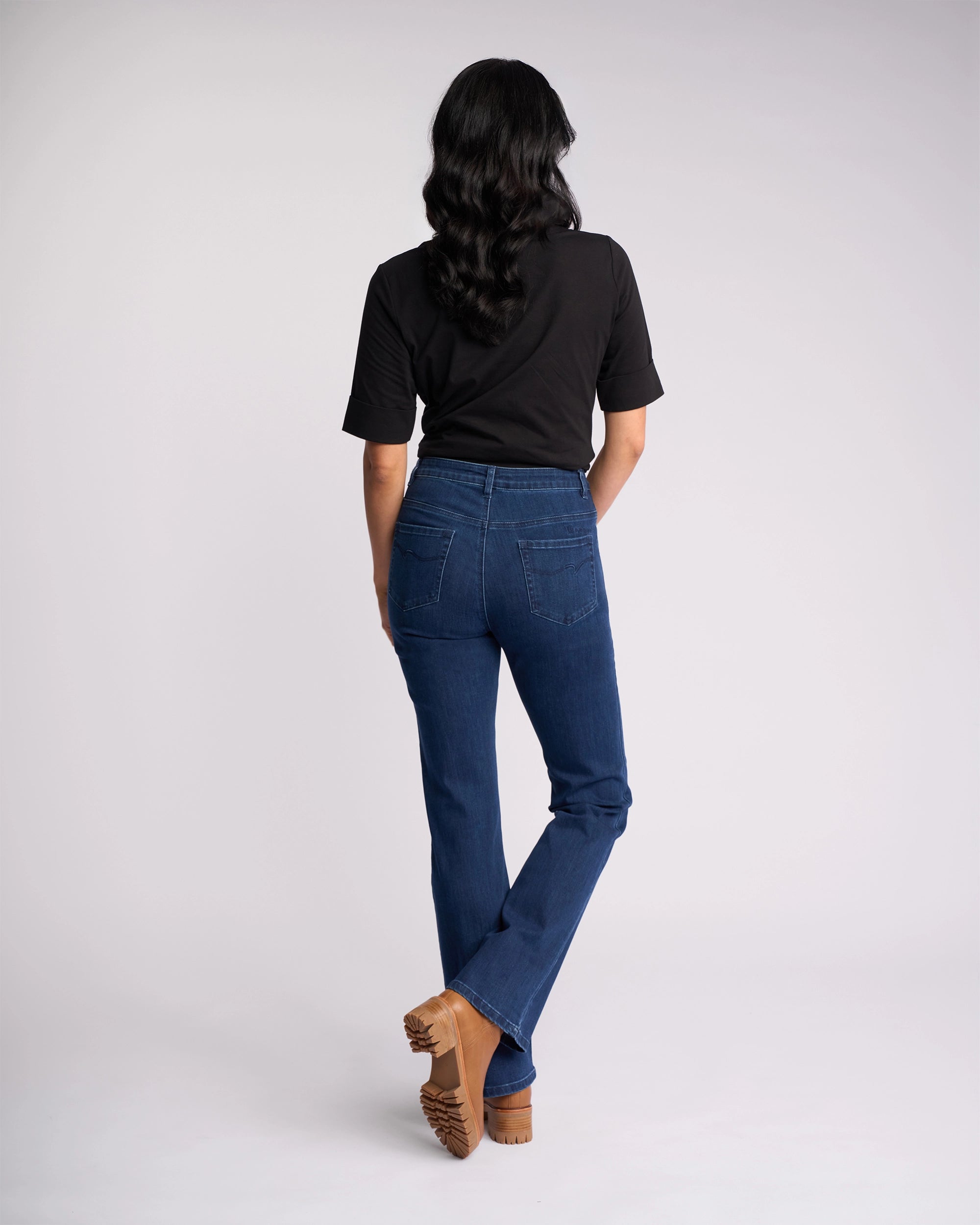 A person with long, wavy hair faces away, wearing a black shirt, brown heeled shoes, and Vassalli’s Theory Jean—Blue Denim (Without Fade)—against a plain light background.