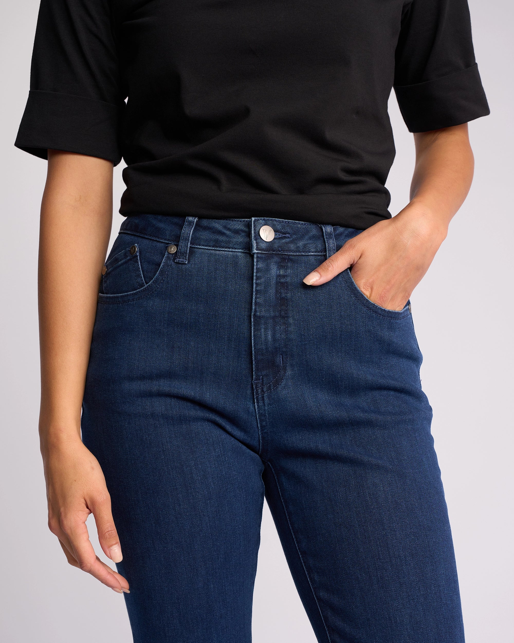 A person wearing a black short-sleeve top and Vassalli Theory Jean - Blue Denim (Without Fade) stands with one hand in a pocket and the other arm relaxed by their side, shown from shoulders to thighs.