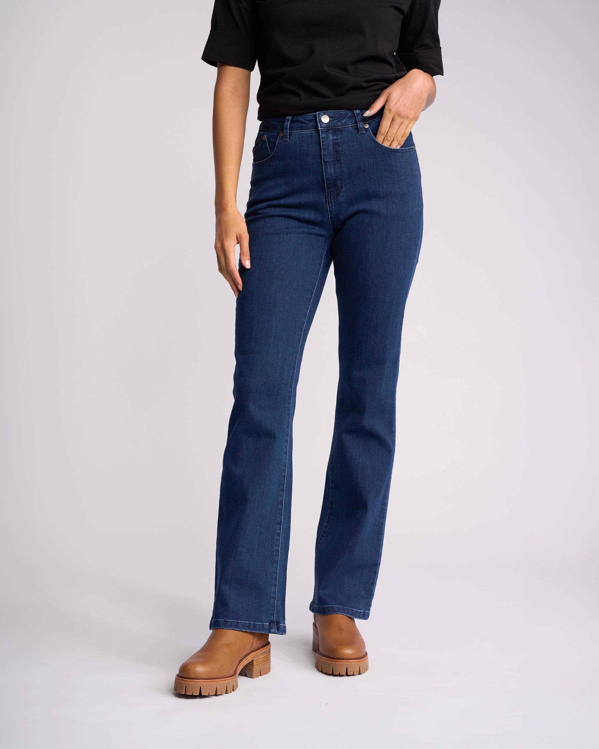A person wearing a black top, Vassalli Theory Jean - Blue Denim (Without Fade), and brown heeled boots stands against a plain light background. The person's face is not visible.