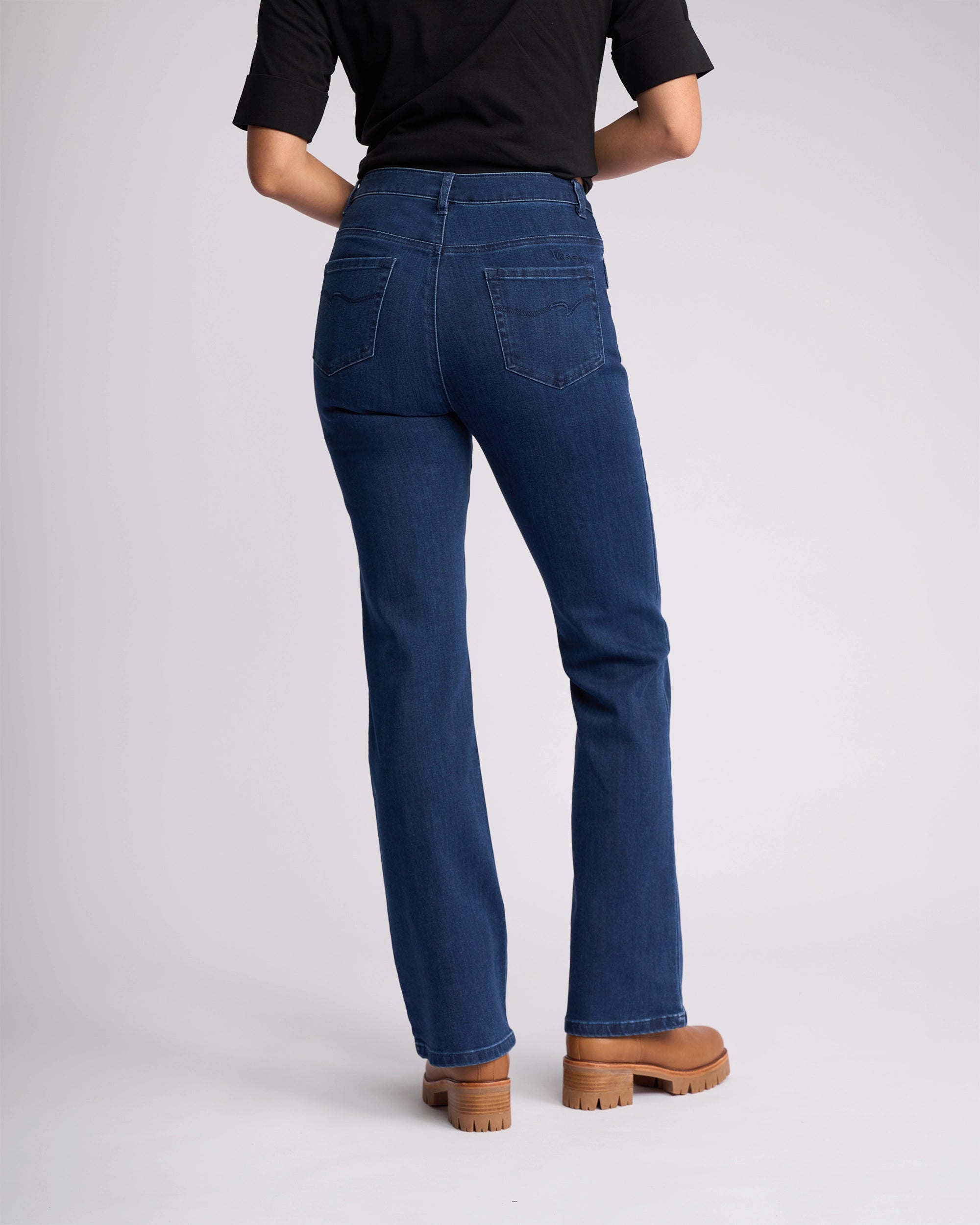 A person faces away, showcasing the back of Vassalli's Theory Jean - Blue Denim (Without Fade), paired with a black top and tan platform boots, against a plain light background.