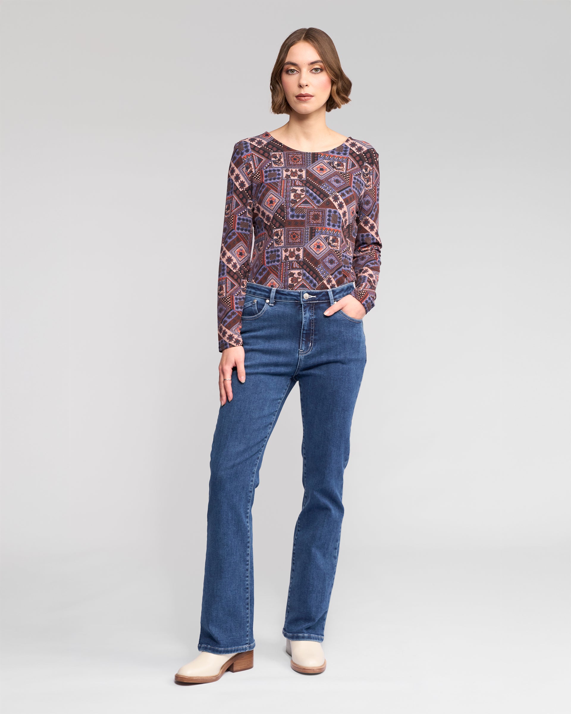 A woman stands against a plain background, wearing Vassalli's Theory Jean in Dark Blue Brushed with a patterned long-sleeve top and light ankle boots, one hand in her pocket.