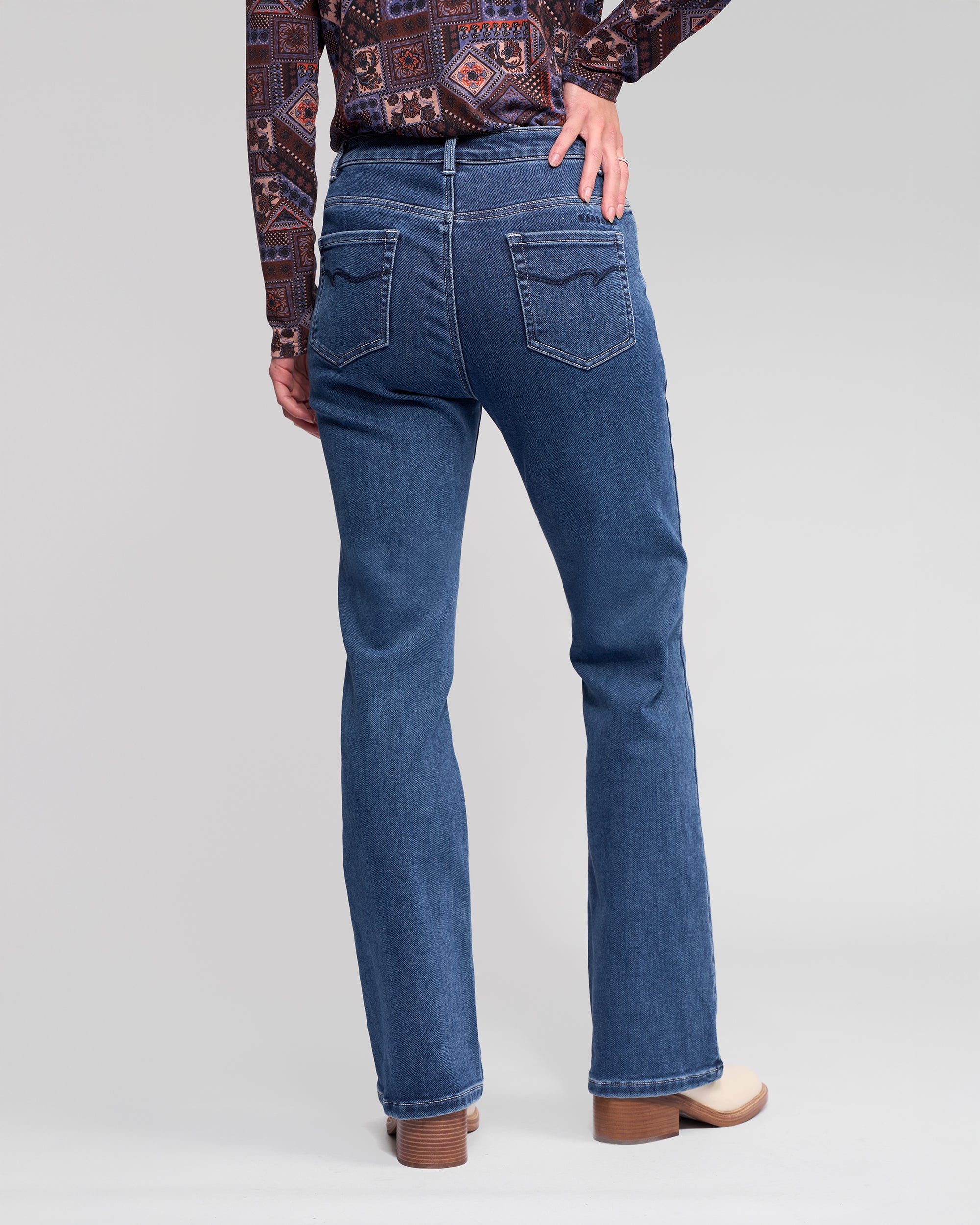 Someone is standing with one hand on their hip, facing away from the camera in Vassalli Theory Jean - Dark Blue Brushed, a patterned long-sleeve shirt, and beige heeled shoes against a plain background.