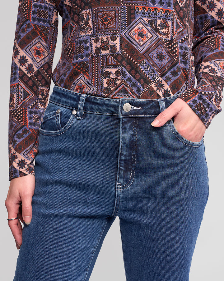 Wearing the Vassalli Theory Jean - Dark Blue Brushed, a person stands against a plain background with one hand in their pocket, paired with a long-sleeve shirt featuring a colorful geometric pattern.