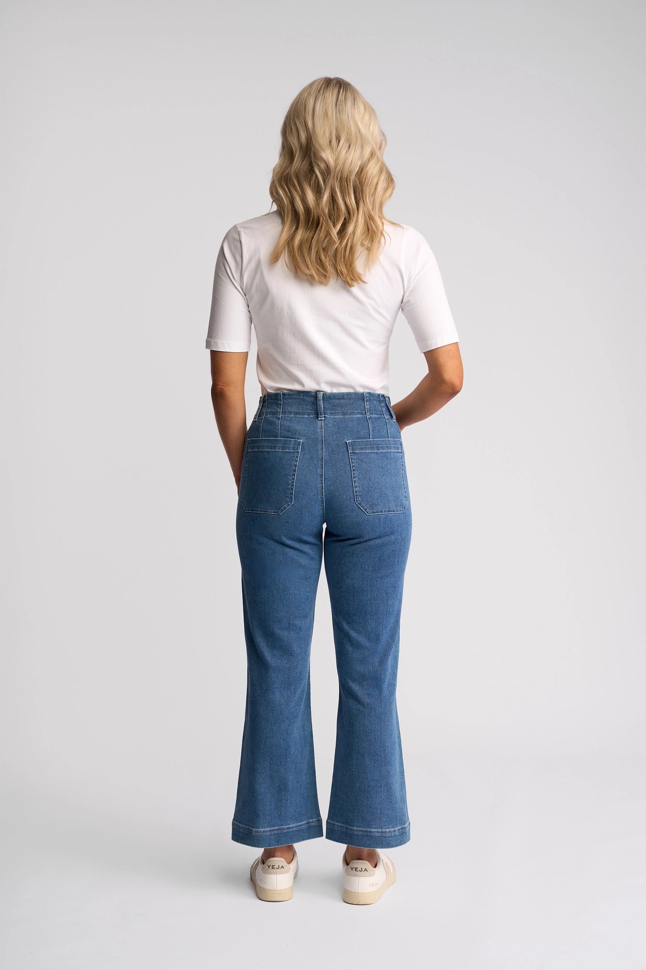 A person with wavy, blonde hair is seen from behind, standing against a plain light background while wearing a white short-sleeve top paired with the Vassalli Daily Jean in Blue Hue Denim.