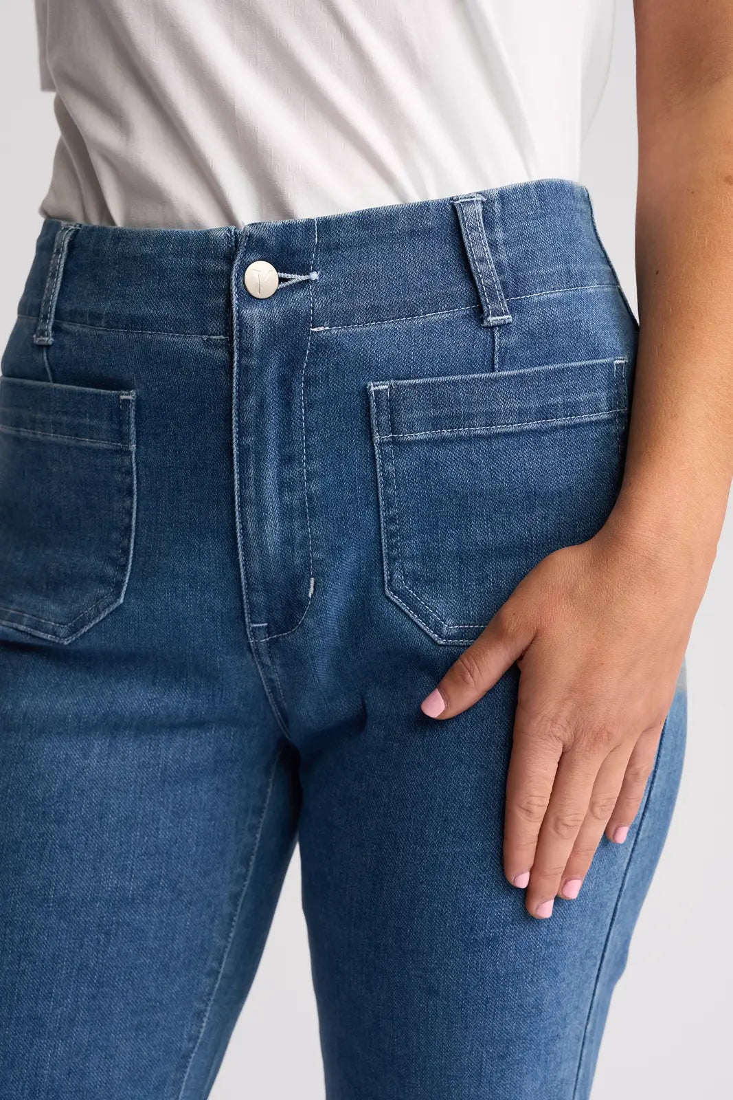Wearing a white shirt and the Vassalli Daily Jean in Blue Hue Denim—high-waisted, straight-leg jeans with front patch pockets—a person rests their right hand on their thigh.