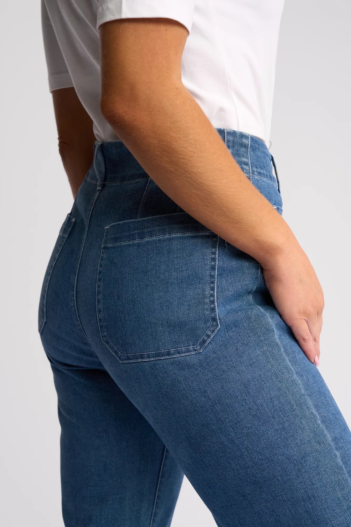 Model wears the Vassalli Daily Jean in Blue Hue Denim, a high-waisted, straight-leg style. Shown from the waist down at an angle, the fit is highlighted as one hand rests near the back pocket alongside a white short-sleeve shirt.