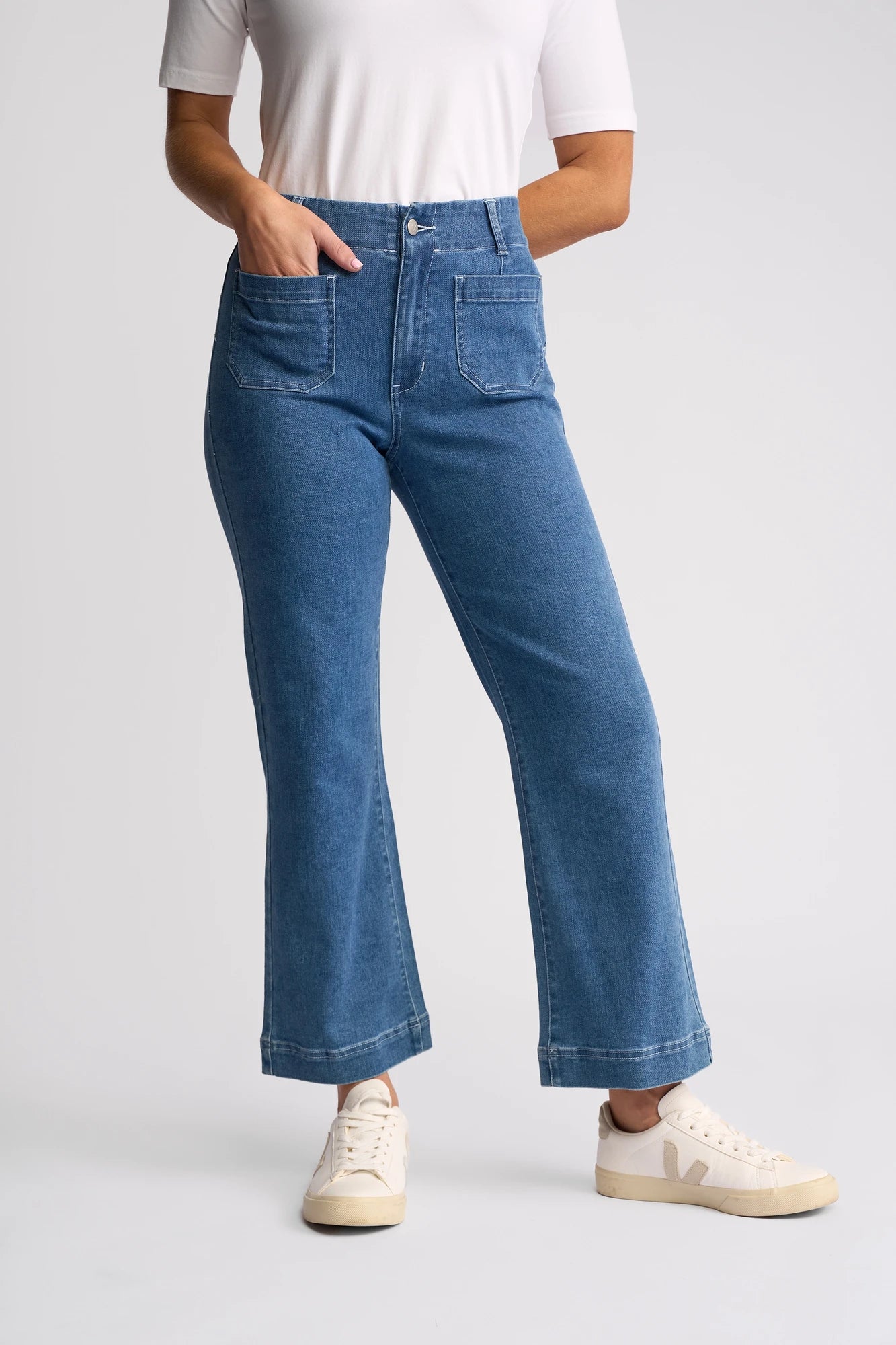 A person is wearing a white top, Vassalli Daily Jean - Blue Hue Denim high-waisted stretch straight-leg jeans with front pockets, and white sneakers, standing against a plain light background.
