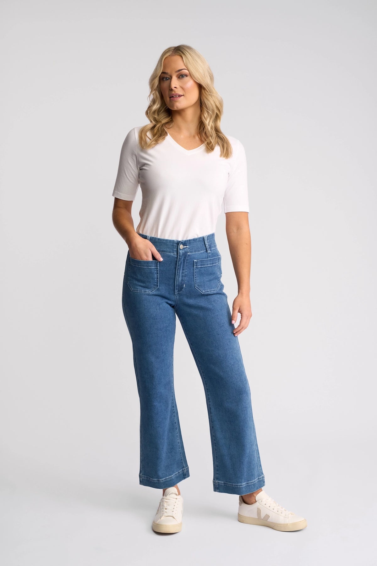 A woman with blonde, wavy hair stands against a plain background, wearing a white short-sleeve top and the Vassalli Daily Jean in Blue Hue Denim. The high-waisted, straight-leg jeans feature front pockets and are made from stretch denim. She completes her look with white sneakers and has her left hand tucked into her pocket.