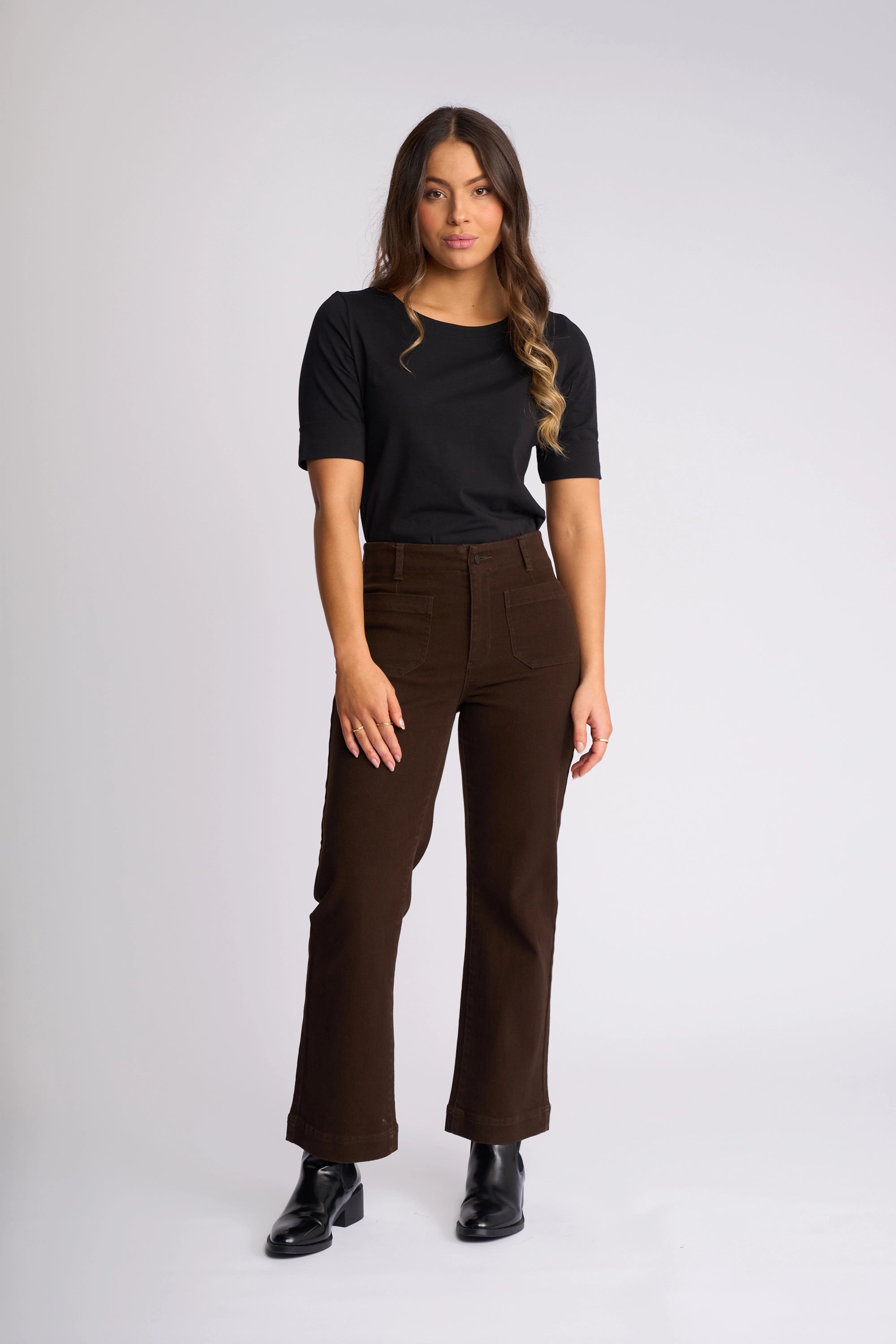 A woman stands against a plain background, wearing the Vassalli Daily Jean in Chocolate—high-waisted, brown stretch denim straight-leg jeans with patch pockets—paired with a black short-sleeve top and black shoes. Her long wavy hair is styled over one shoulder.
