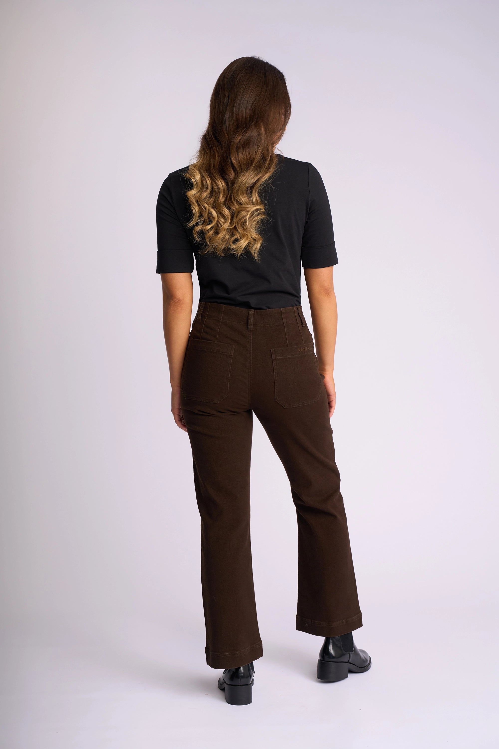A person with long wavy brown hair, wearing a black shirt, Vassalli Daily Jean - Chocolate stretch denim straight-leg jeans with patch pockets, and black shoes, stands facing away from the camera against a plain light background.