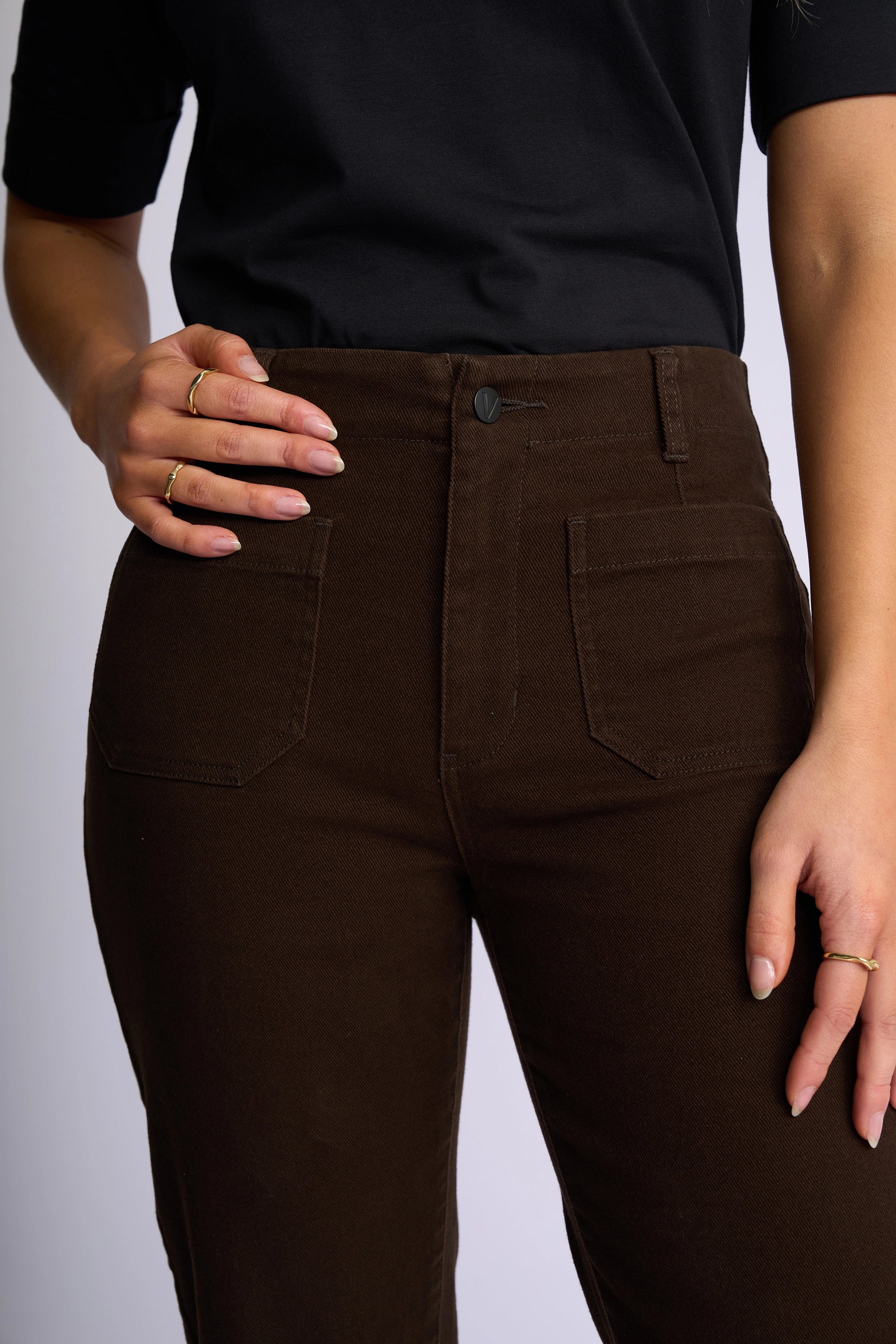A person wearing a black short-sleeve top and the Vassalli Daily Jean in Chocolate, featuring high-waisted stretch denim with patch pockets. One hand rests on their waistband while the other hangs relaxed by their side, and they accessorize with multiple gold rings.