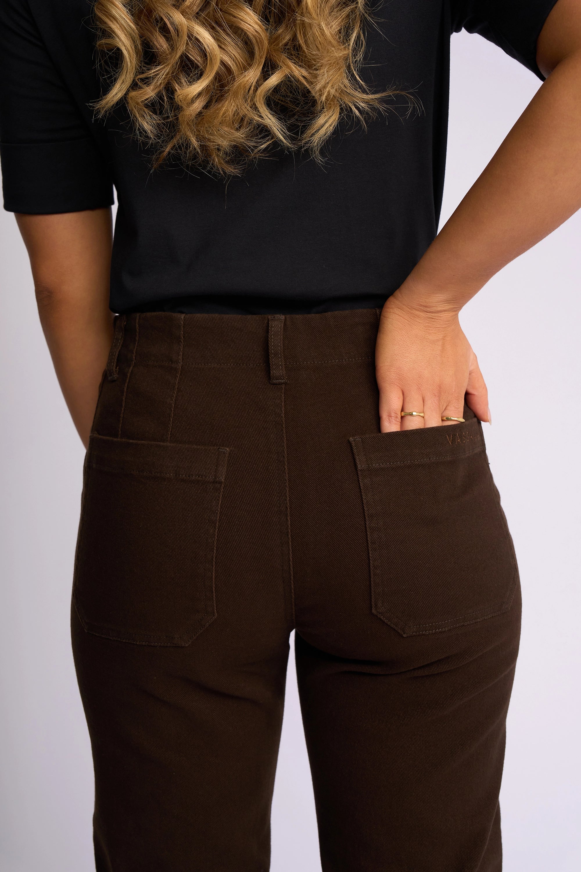 A person is shown from the back, waist to upper arms visible, standing with one hand in the patch pocket of their Vassalli Daily Jean in Chocolate. They are wearing a black shirt and brown pants with a straight-leg fit.