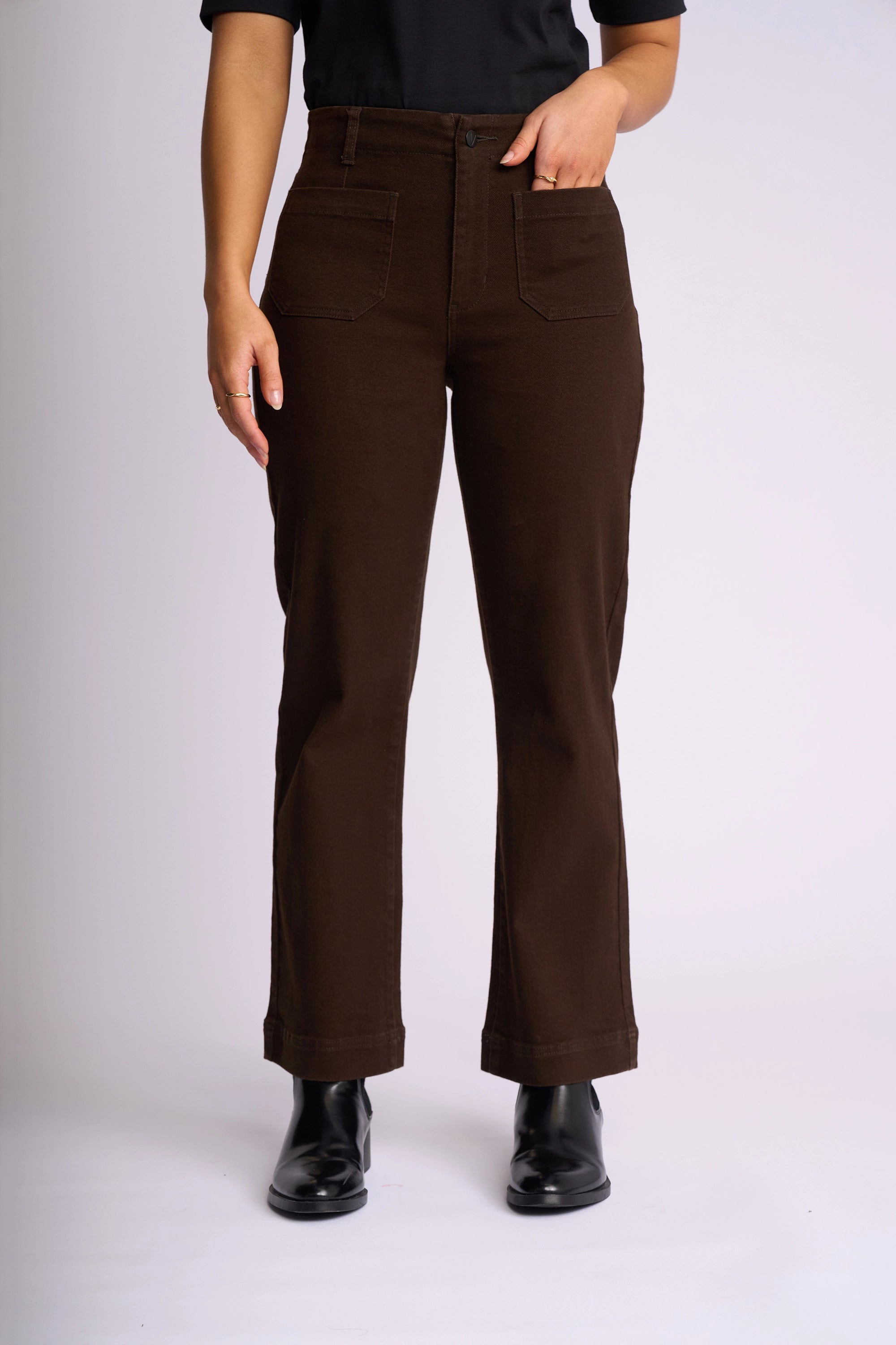 Wearing the Vassalli Daily Jean in Chocolate, styled with black boots and a black shirt, this person stands against a plain light background with one hand casually tucked into their pocket.