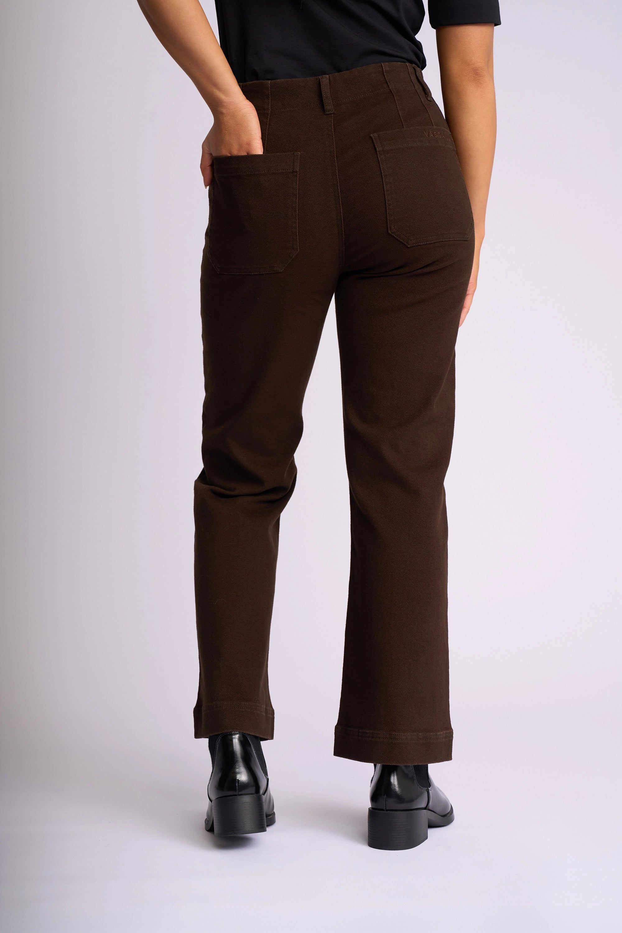 A person stands against a plain background, facing away. They wear a black shirt, Vassalli's Daily Jean in Chocolate with patch pockets at the back, and black ankle boots. One hand is placed in a pocket.