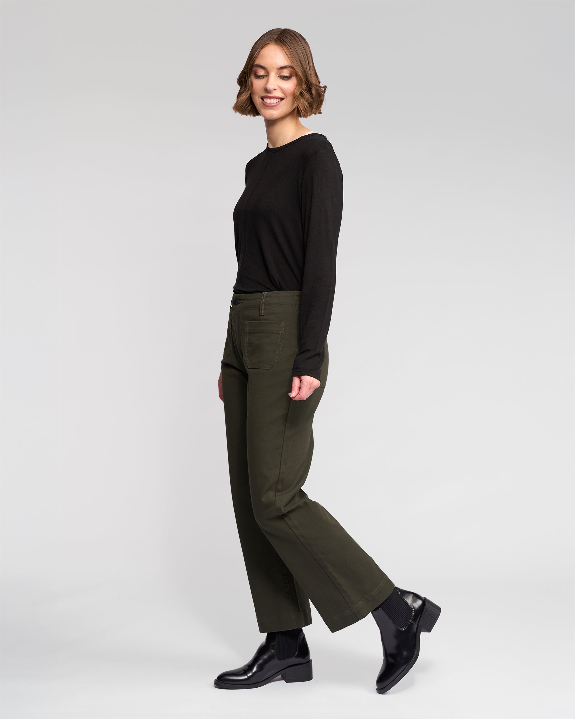 A woman with short brown hair wears the Vassalli Daily Jean - Hunter and a black long-sleeve shirt with black ankle boots. She stands sideways, smiling, against a plain white background.