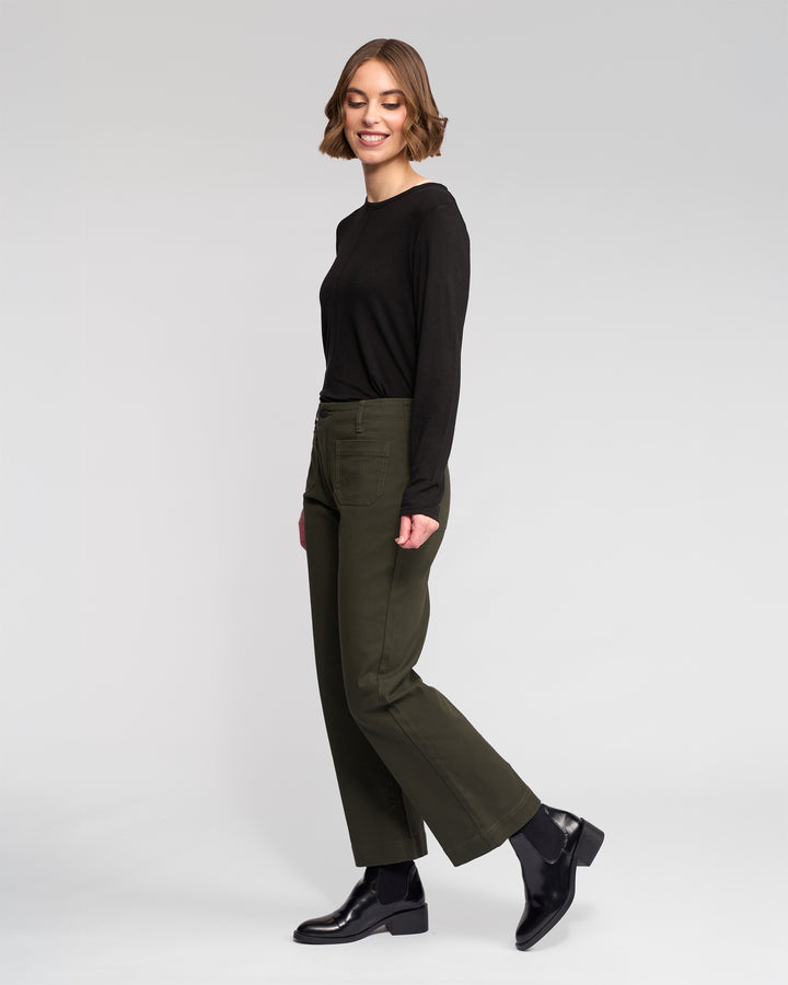 A woman with short brown hair wears the Vassalli Daily Jean - Hunter and a black long-sleeve shirt with black ankle boots. She stands sideways, smiling, against a plain white background.
