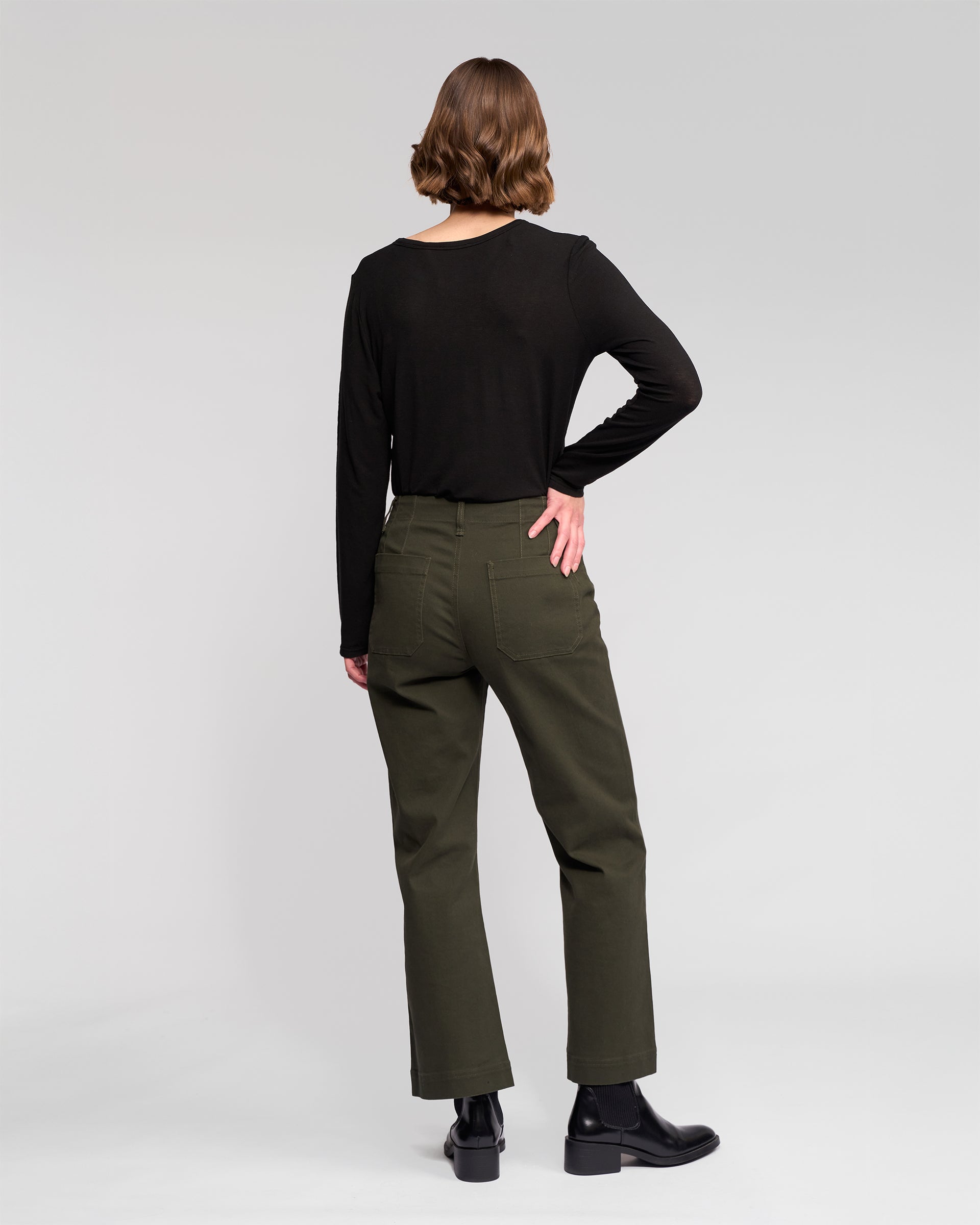 A person with short brown hair, in a long-sleeve black top, Vassalli’s Daily Jean - Hunter, and black boots stands facing away from the camera against a plain light background.