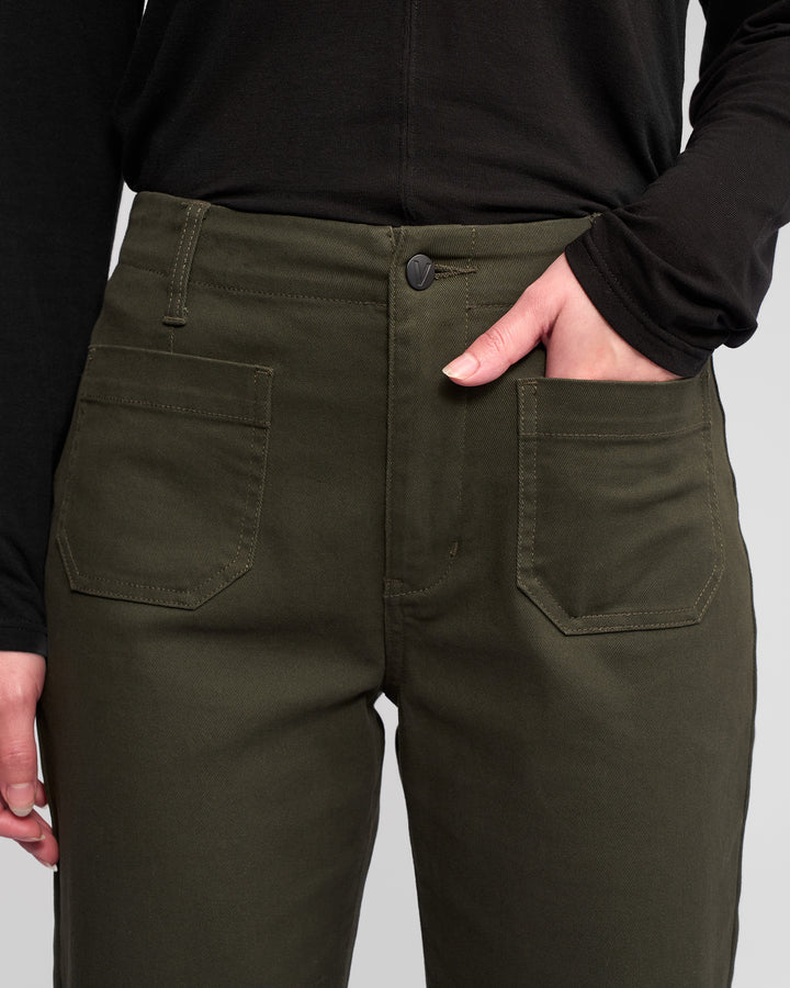 A person wears Vassalli's Daily Jean - Hunter, dark green ankle-grazer pants with front patch pockets, paired with a black long-sleeve top and one hand in the right pocket. The image highlights the pants from waist to mid-thigh.