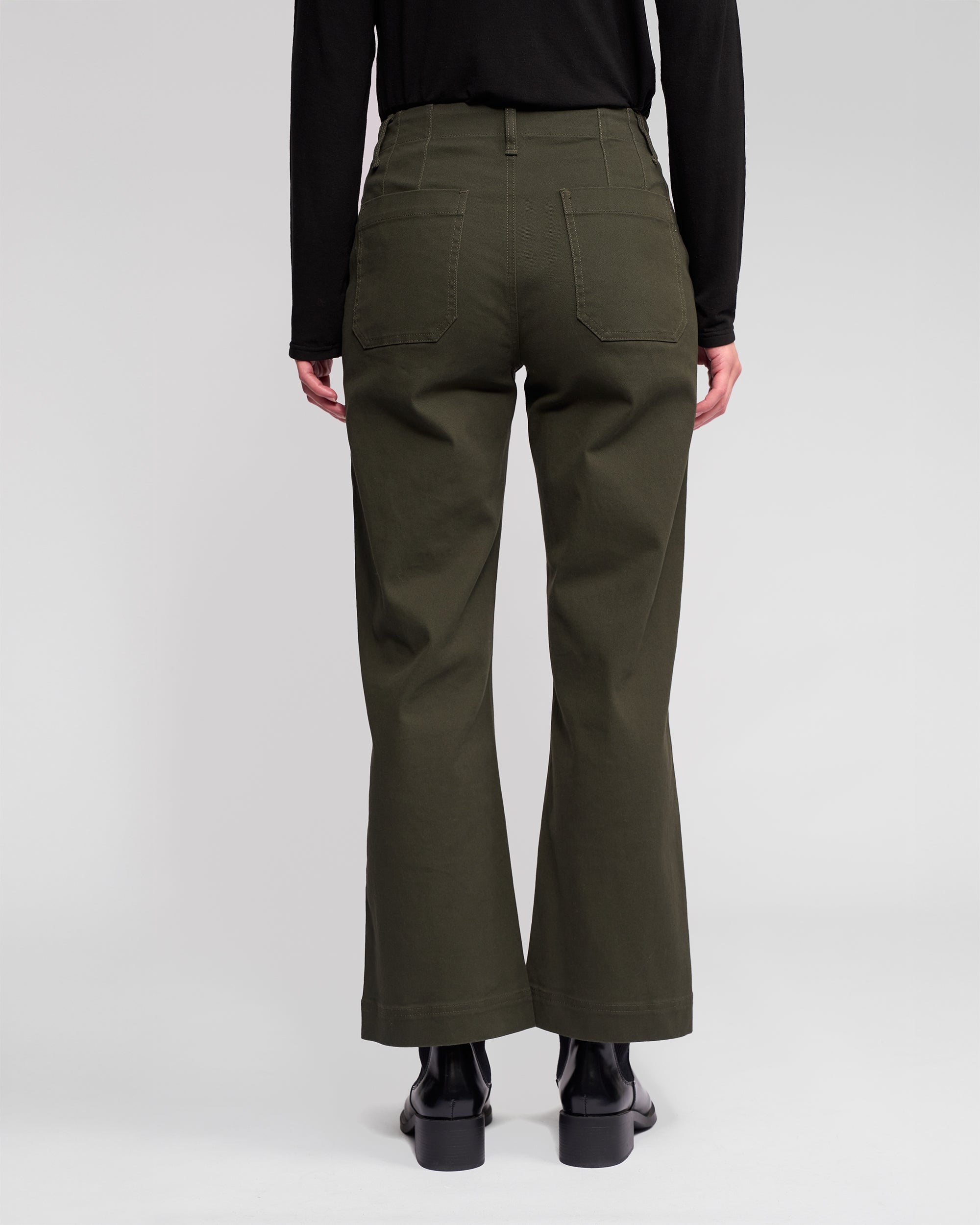 Someone stands with their back to the camera against a light background, dressed in Vassalli's Daily Jean - Hunter in dark green, paired with a black long-sleeve top and black boots.