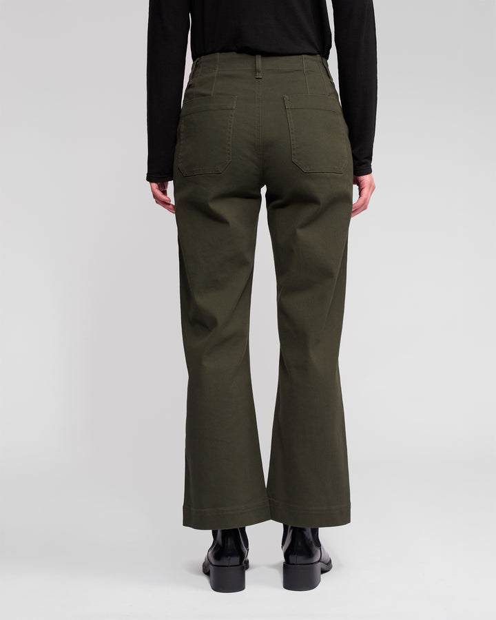 Someone stands with their back to the camera against a light background, dressed in Vassalli's Daily Jean - Hunter in dark green, paired with a black long-sleeve top and black boots.