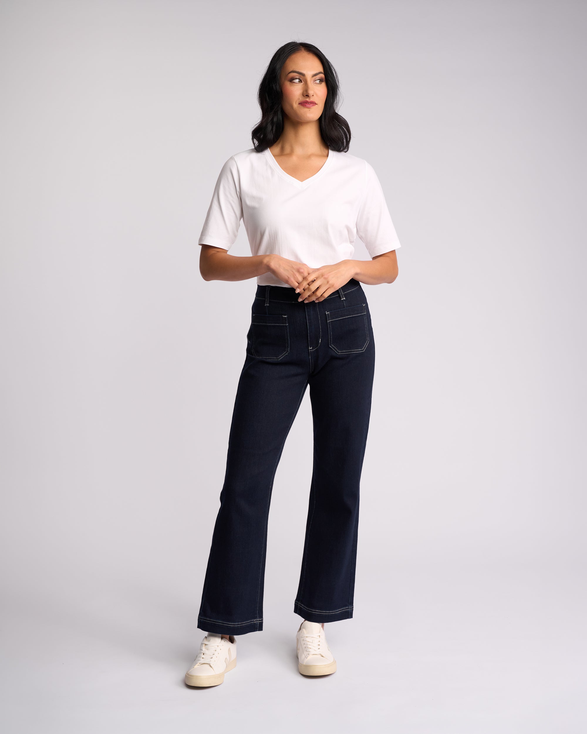 A woman stands against a light background, wearing Vassalli’s Daily Jean - Ink Denim: high-waisted, dark blue straight leg jeans with stretch and front pockets, paired with a white V-neck t-shirt and white sneakers. She looks slightly to the side.