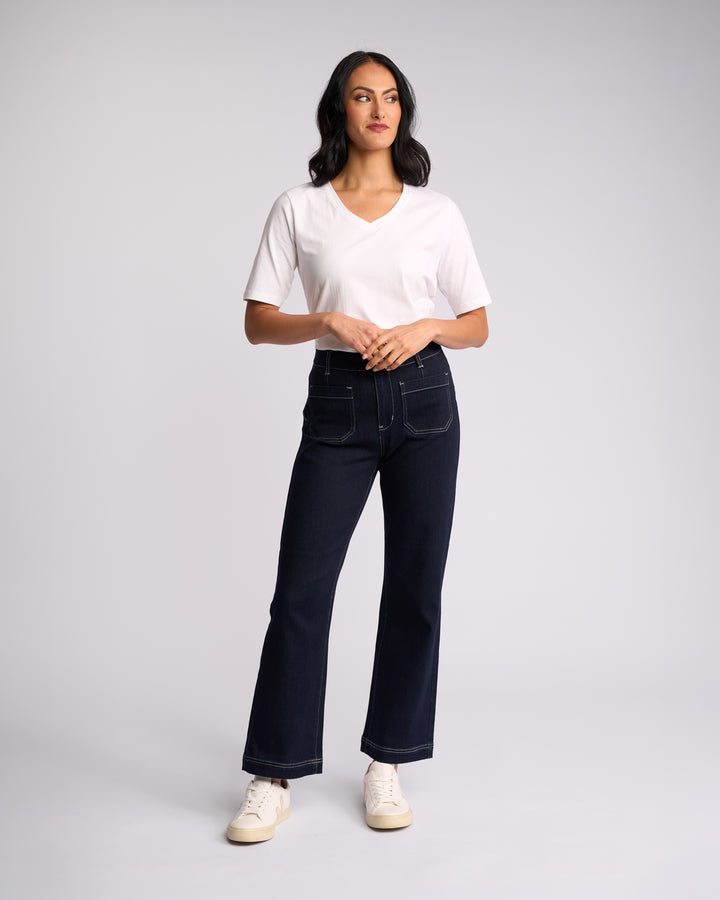 A woman stands against a light background, wearing Vassalli’s Daily Jean - Ink Denim: high-waisted, dark blue straight leg jeans with stretch and front pockets, paired with a white V-neck t-shirt and white sneakers. She looks slightly to the side.