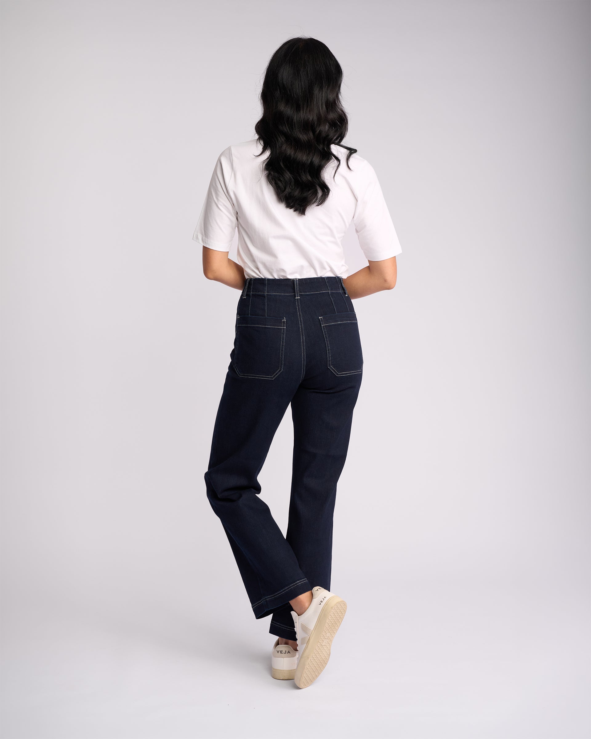 A person with long dark hair, wearing a white t-shirt, Vassalli Daily Jean in Ink Denim, and white sneakers stands facing away from the camera against a plain light background.