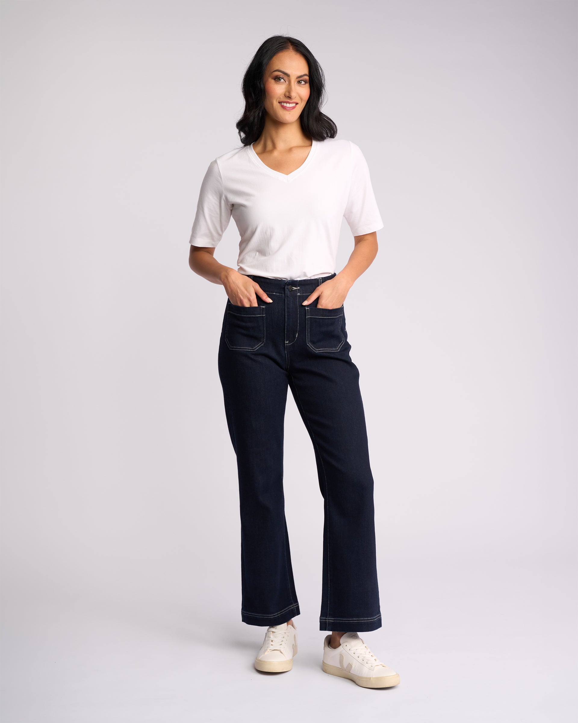 A person stands against a plain background, smiling with hands in pockets, wearing a white V-neck T-shirt, white sneakers, and Vassalli Daily Jean - Ink Denim: dark high-waisted straight leg jeans with front pockets in stretch fabric.