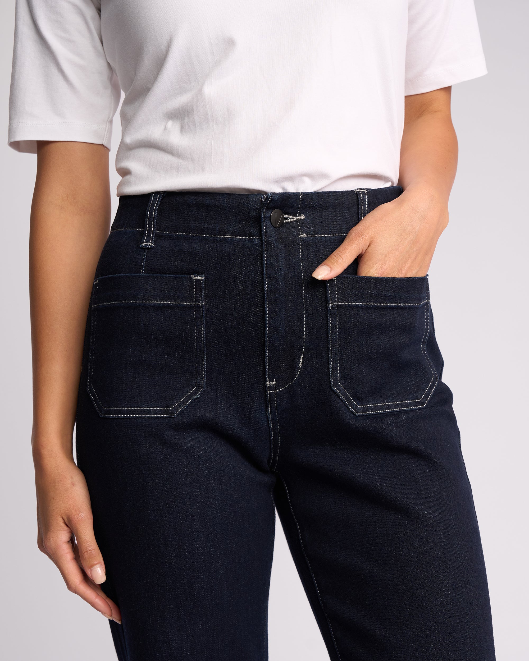 A person stands against a neutral background wearing a white short-sleeve shirt and Vassalli’s Daily Jean - Ink Denim, high-waisted dark blue straight leg jeans with white stitching, with one hand in the front pocket.