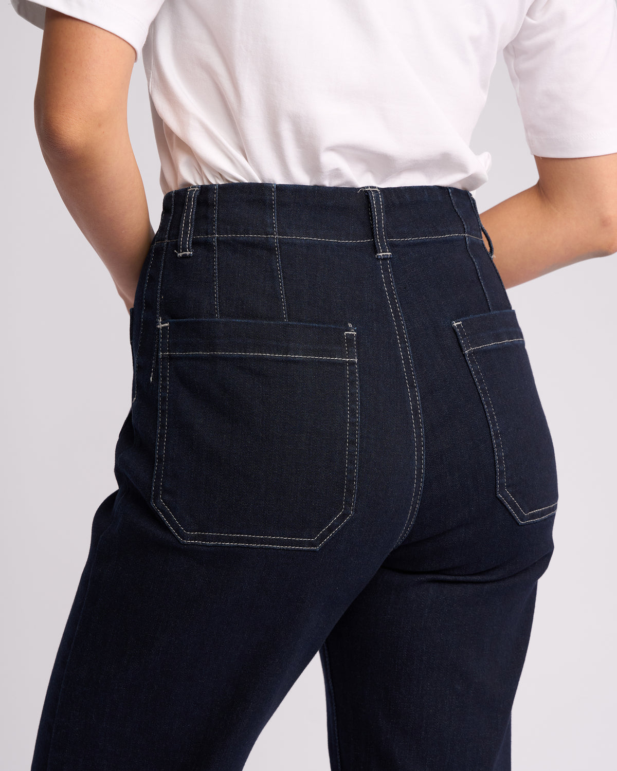 A person wearing Vassalli Daily Jean - Ink Denim, high-waisted dark blue straight leg jeans with white stitching, and a white T-shirt, shown from the waist down and viewed from behind.