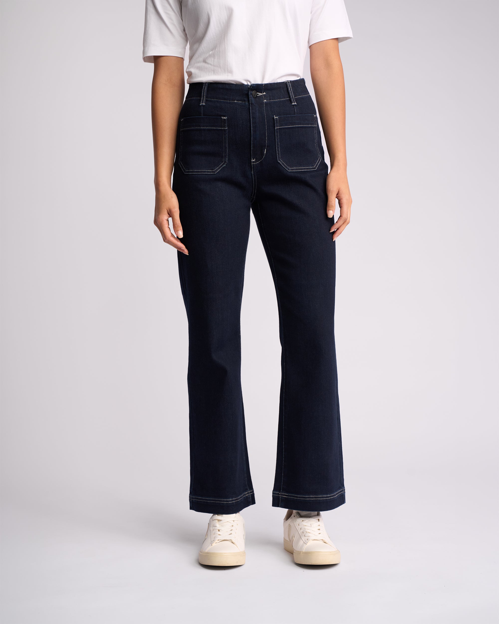 A person stands against a plain background in a white short-sleeve shirt, Vassalli Daily Jean - Ink Denim (dark blue, ankle grazer with front pockets, stretch fabric), and white sneakers. The head is not visible.
