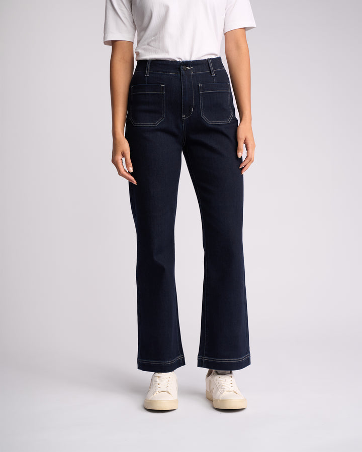 A person stands against a plain background in a white short-sleeve shirt, Vassalli Daily Jean - Ink Denim (dark blue, ankle grazer with front pockets, stretch fabric), and white sneakers. The head is not visible.