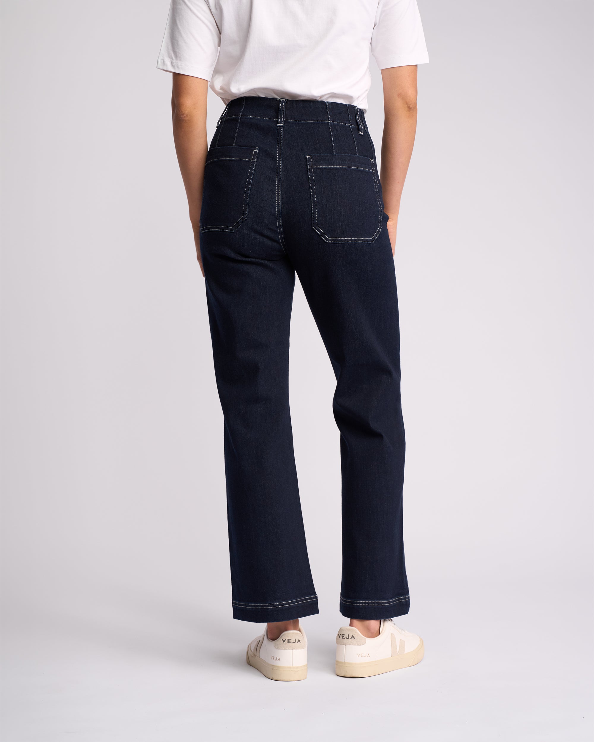 Person facing away, wearing a white t-shirt and Vassalli Daily Jean - Ink Denim, featuring dark high-waisted straight leg fit with visible stitching and stretch fabric, paired with white Veja sneakers against a light background.