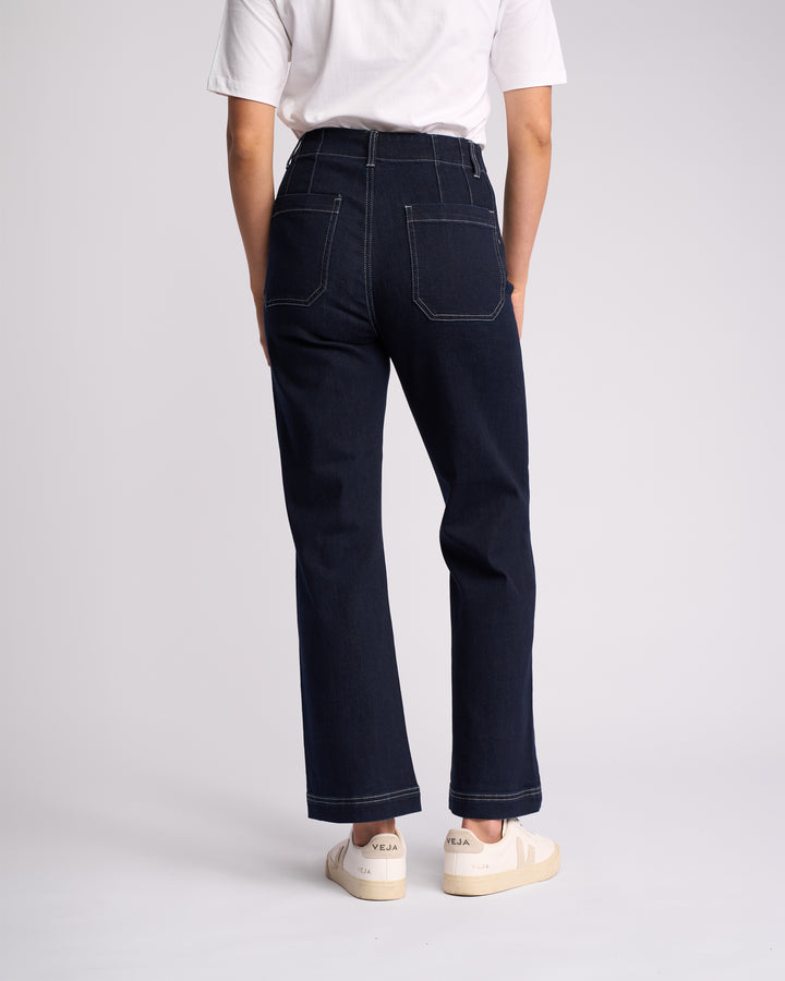 Person facing away, wearing a white t-shirt and Vassalli Daily Jean - Ink Denim, featuring dark high-waisted straight leg fit with visible stitching and stretch fabric, paired with white Veja sneakers against a light background.