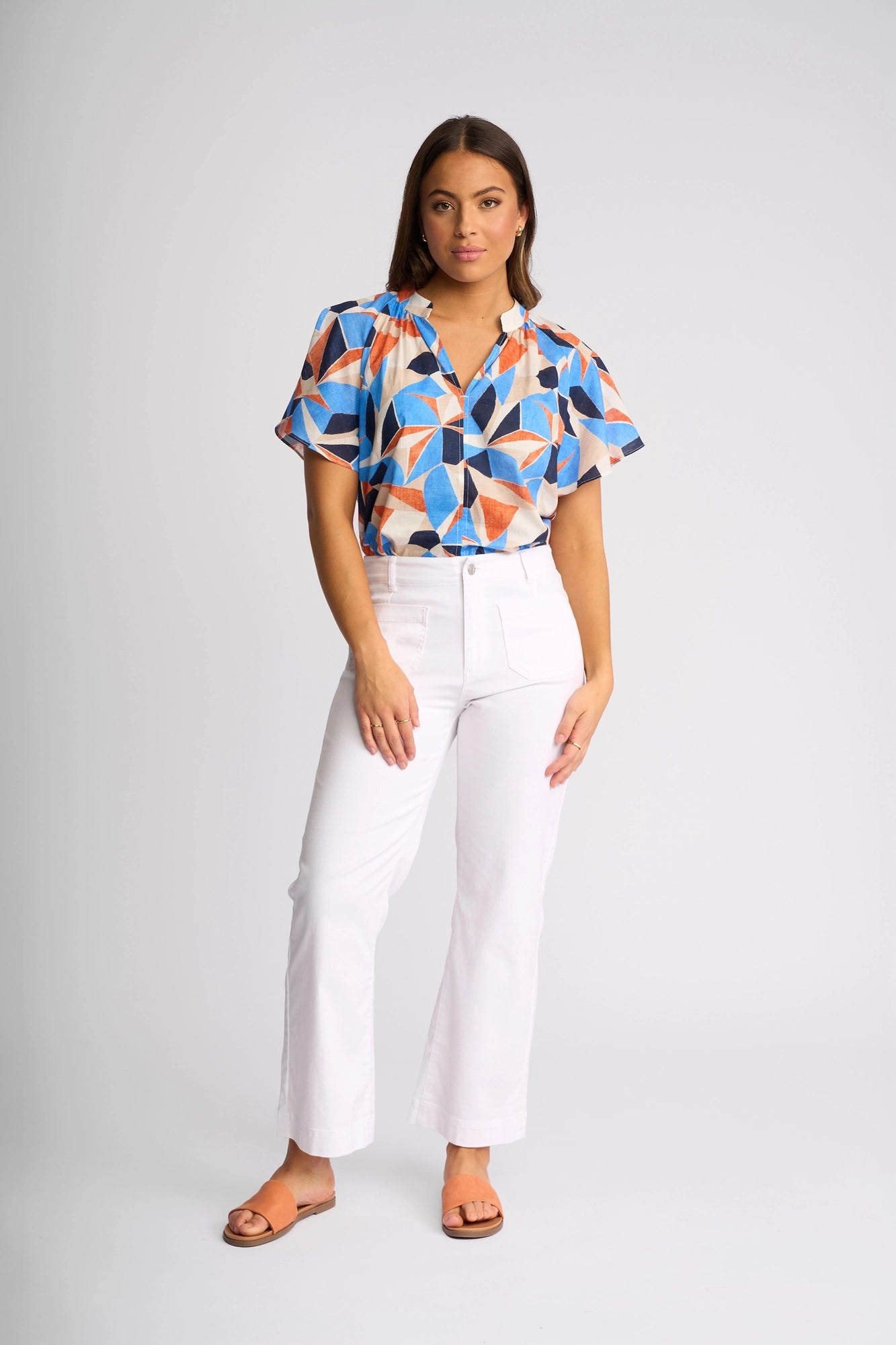 A woman stands facing forward, wearing a colorful geometric-patterned blouse, the Vassalli Daily Jean in White Denim with an ankle-grazer cut, and brown sandals. She has long brown hair and a neutral expression. The background is plain light gray.