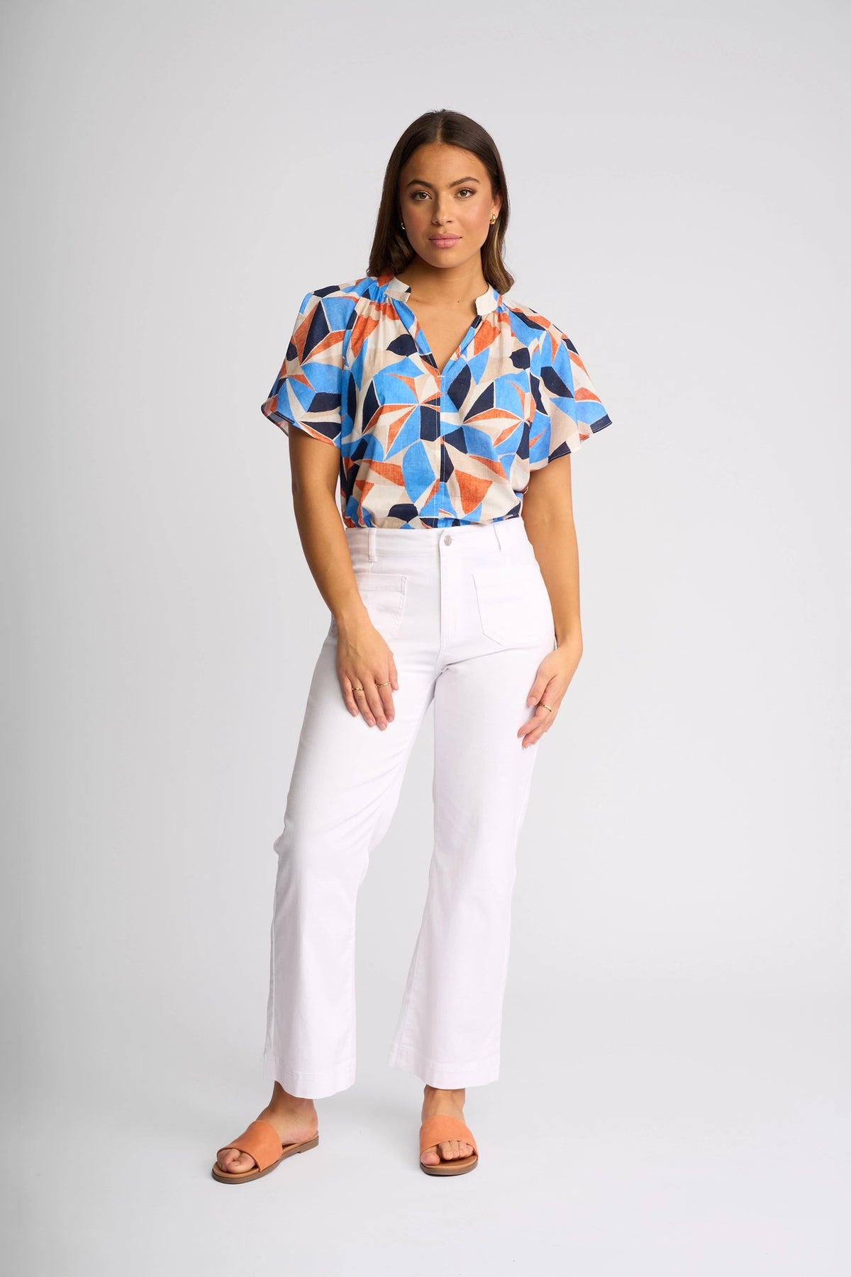 A woman stands facing forward, wearing a colorful geometric-patterned blouse, the Vassalli Daily Jean in White Denim with an ankle-grazer cut, and brown sandals. She has long brown hair and a neutral expression. The background is plain light gray.