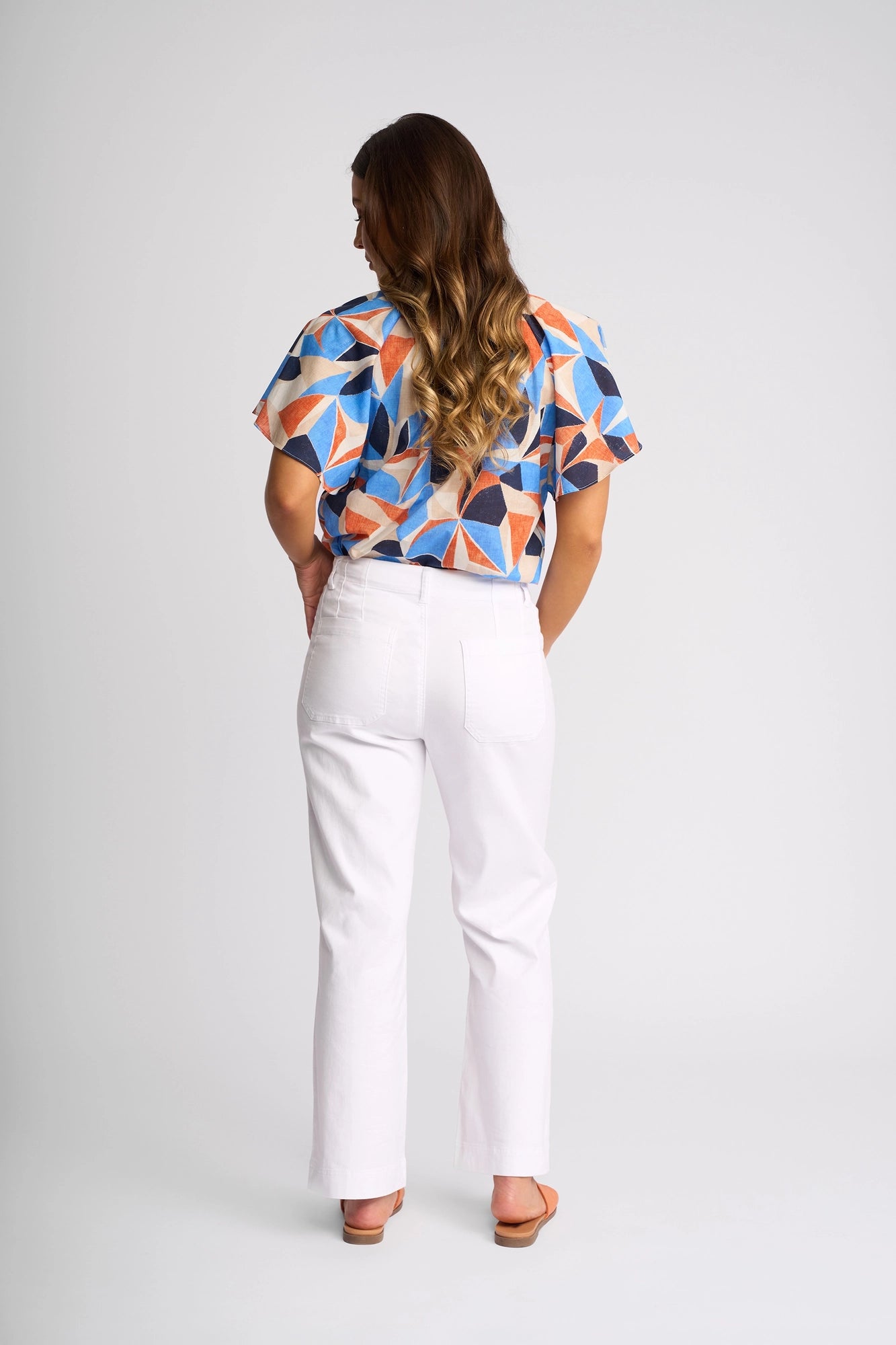 A woman stands facing away, wearing the Vassalli Daily Jean in White Denim paired with a colorful geometric-patterned top. Her long wavy hair cascades down her back as she poses against a plain, light background.