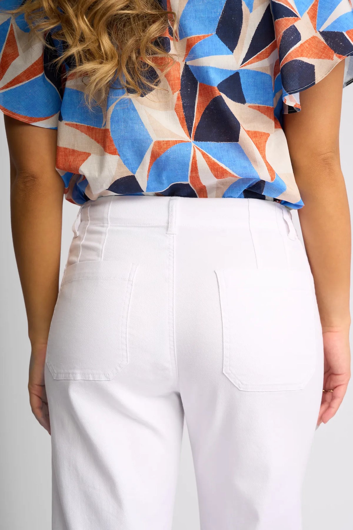 A person stands facing away, dressed in high-waisted Vassalli Daily Jean - White Denim with back pockets, styled with a blue, white, and orange geometric-patterned shirt. Their wavy hair cascades over their shoulder, while the straight-leg silhouette of the jeans gives a modern finish to the outfit.