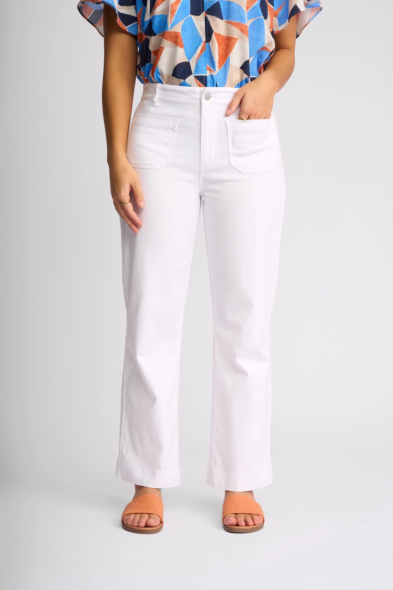 A person models the Vassalli Daily Jean in white denim, paired with a vibrant geometric short-sleeve top and brown sandals, standing against a plain light gray background with their left hand tucked in their pocket.
