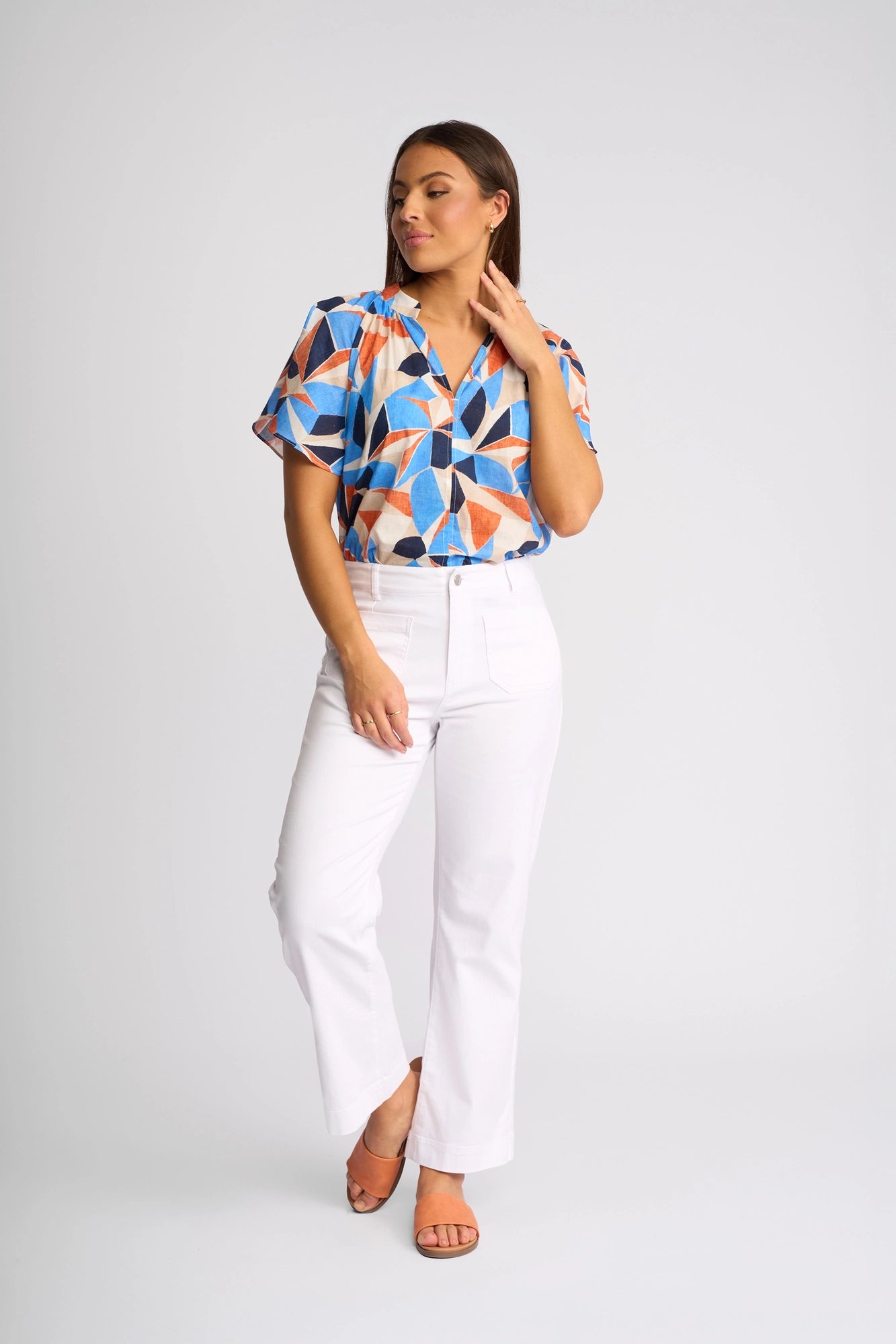 A woman poses against a plain white background wearing the Vassalli Daily Jean in White Denim, styled with a short-sleeve, geometric-patterned blouse featuring blue, orange, and white shapes, and tan heels.
