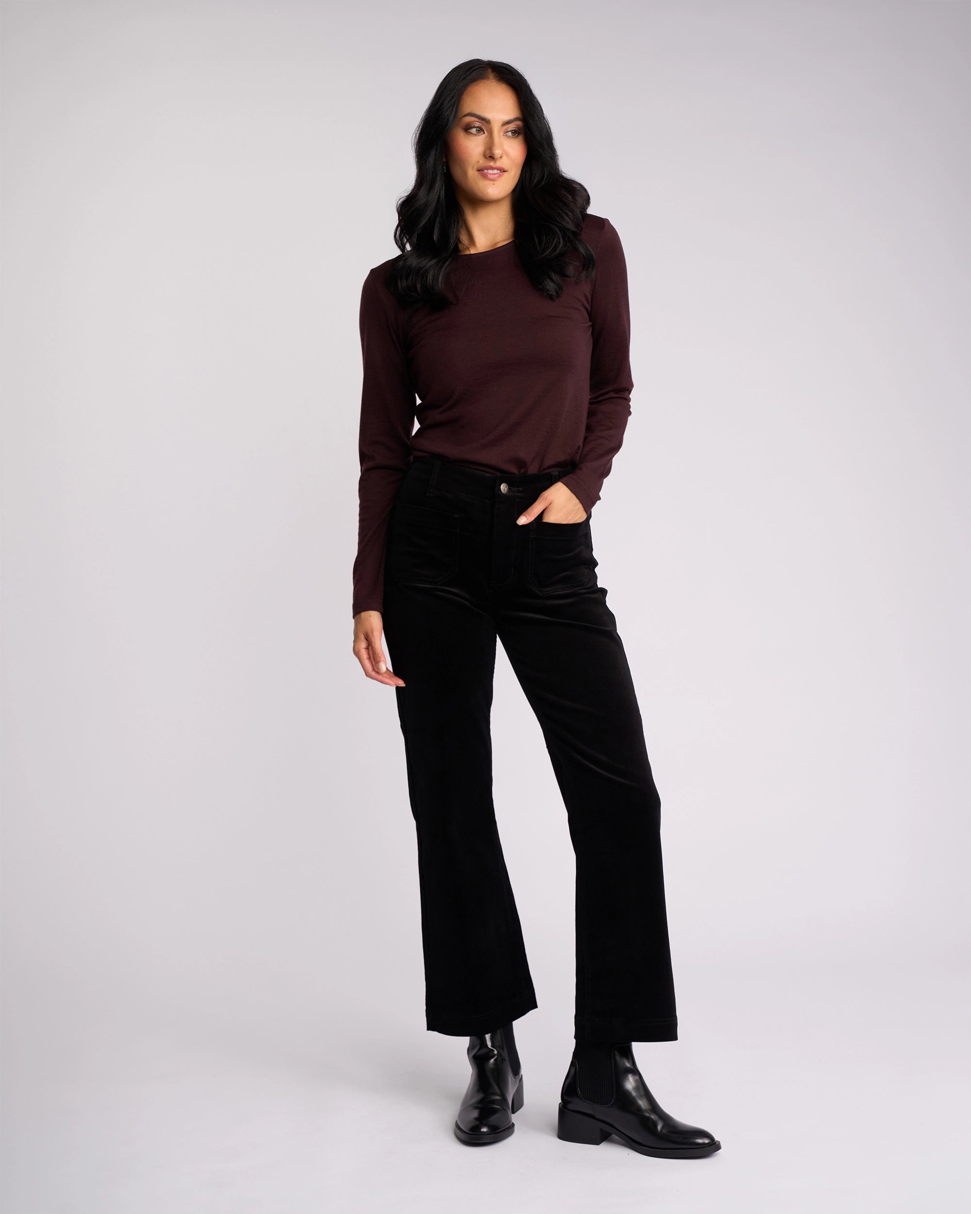 A woman with long dark hair wears the Vassalli Daily Cord Pant in black, paired with a maroon long-sleeve top and black ankle boots. She stands against a light background, one hand in her pocket, the other relaxed by her side.