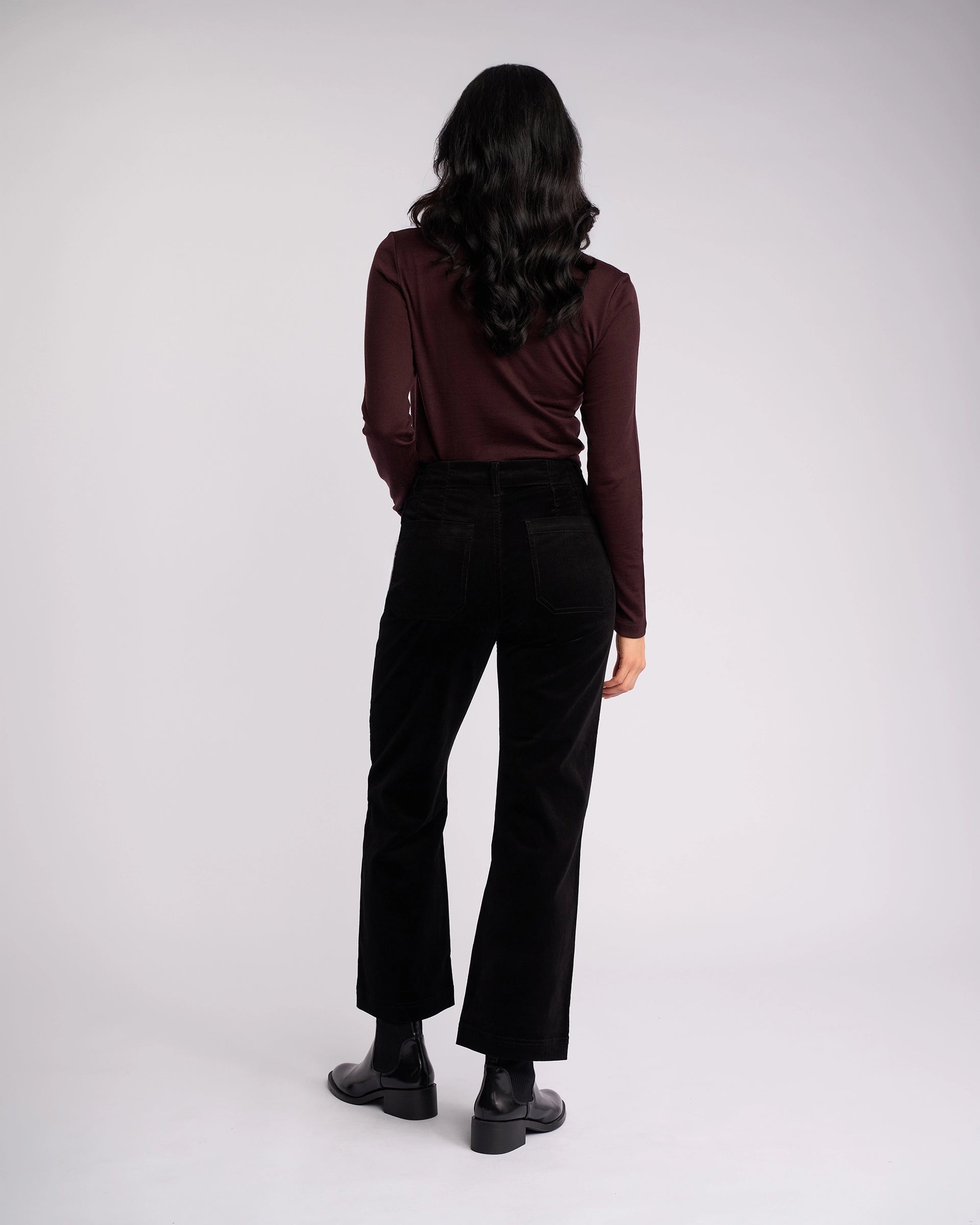 A person with long dark wavy hair stands facing away, wearing Vassalli's Daily Cord Pant in black, a long-sleeve dark maroon top, and black ankle boots against a plain light background.