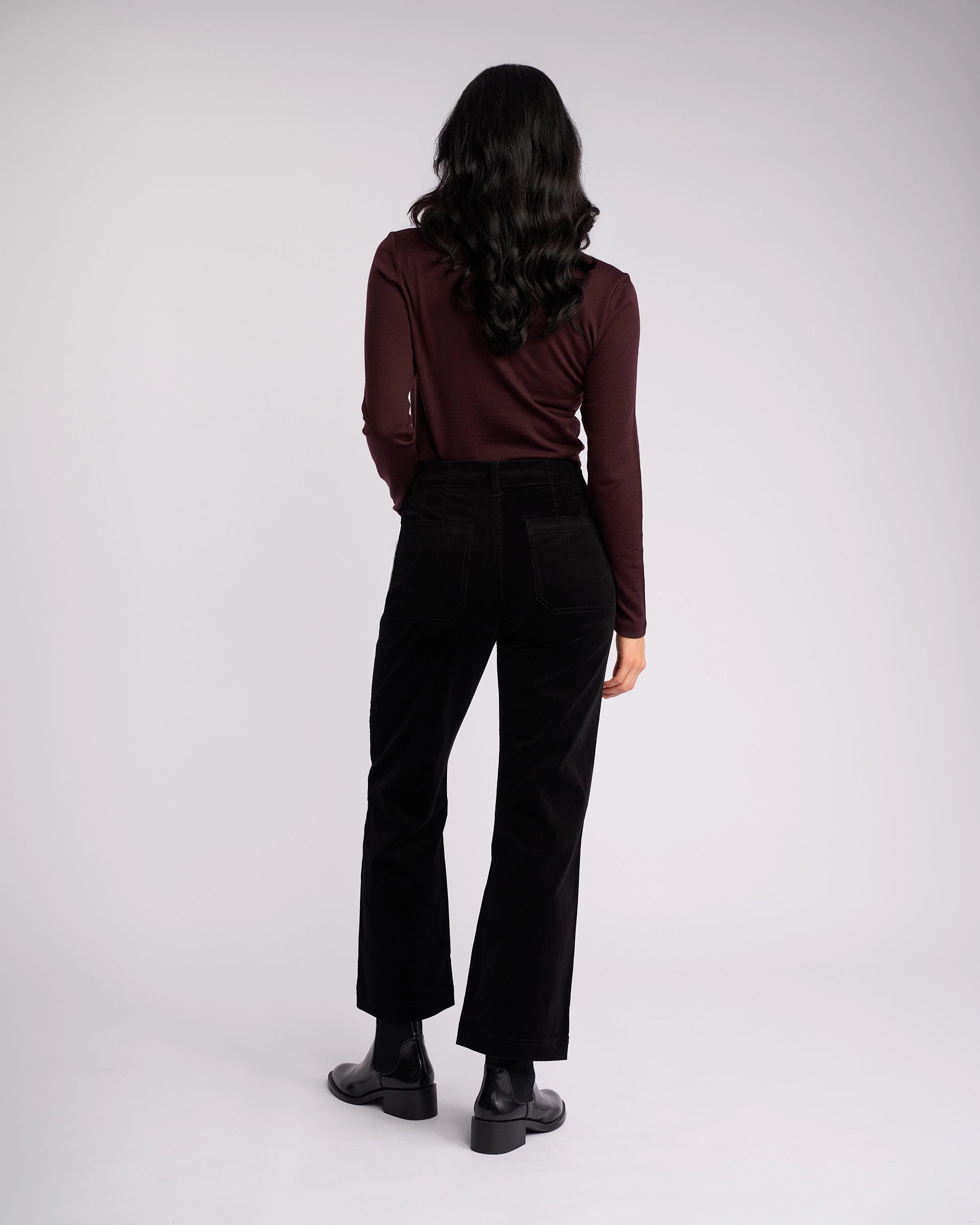 A person with long dark wavy hair stands facing away, wearing Vassalli's Daily Cord Pant in black, a long-sleeve dark maroon top, and black ankle boots against a plain light background.