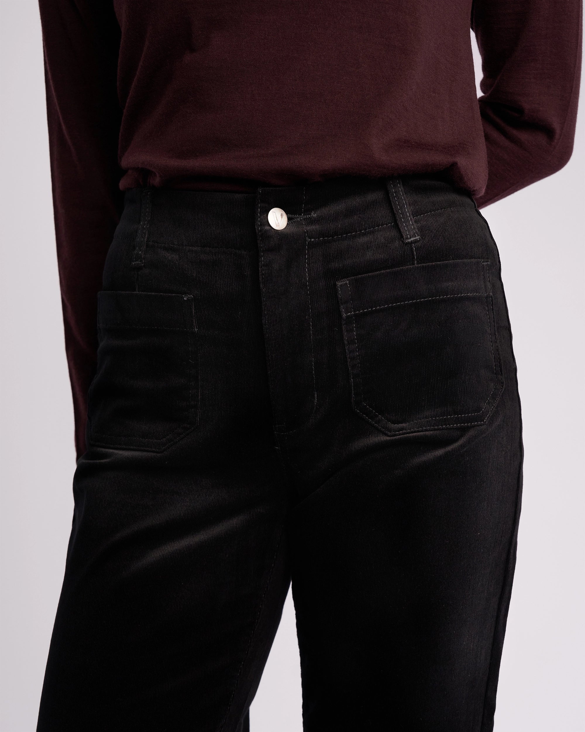 A person wears the Vassalli Daily Cord Pant in black—high-waisted, straight-leg cord pants with front pockets—paired with a dark burgundy long-sleeve shirt, shown from shoulders to upper thighs.